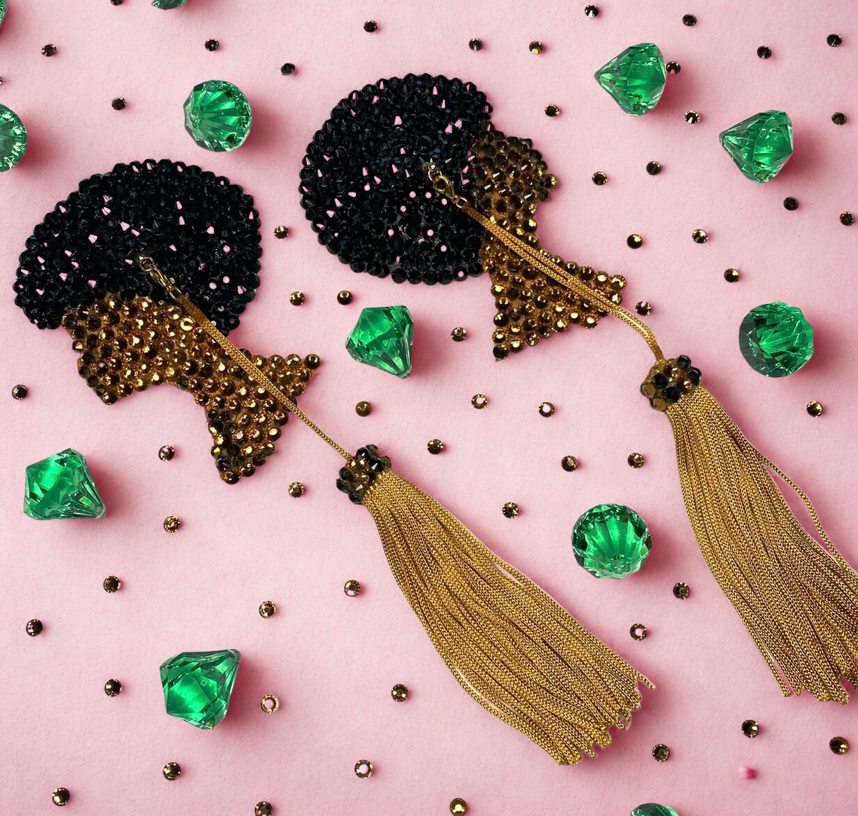 A pair of black and gold sequin tassel earrings are shown on a pink background with green gem shapes and gold beads, echoing the vibrant style of Afro Girl Pasties by Viva La Bombshell Burlesque.