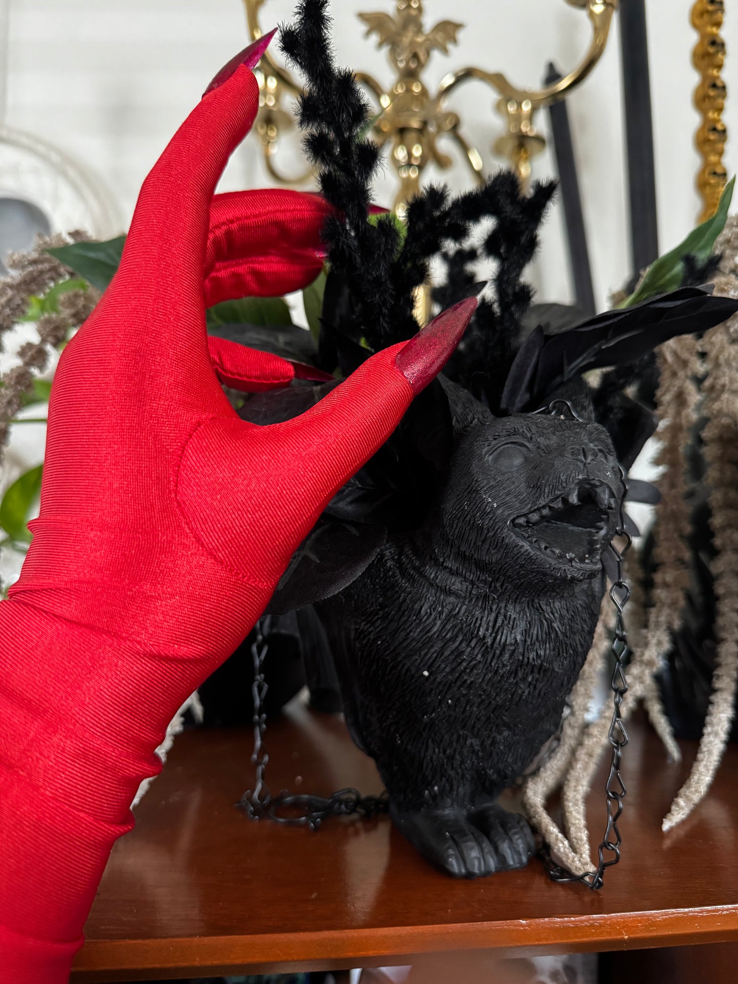 A hand wearing Viva La Bombshell Burlesque's DIY Kitty Claw Gloves with long, red stiletto nails reaches toward a black gargoyle figurine with feathers and chains, surrounded by gold decor and plants.
