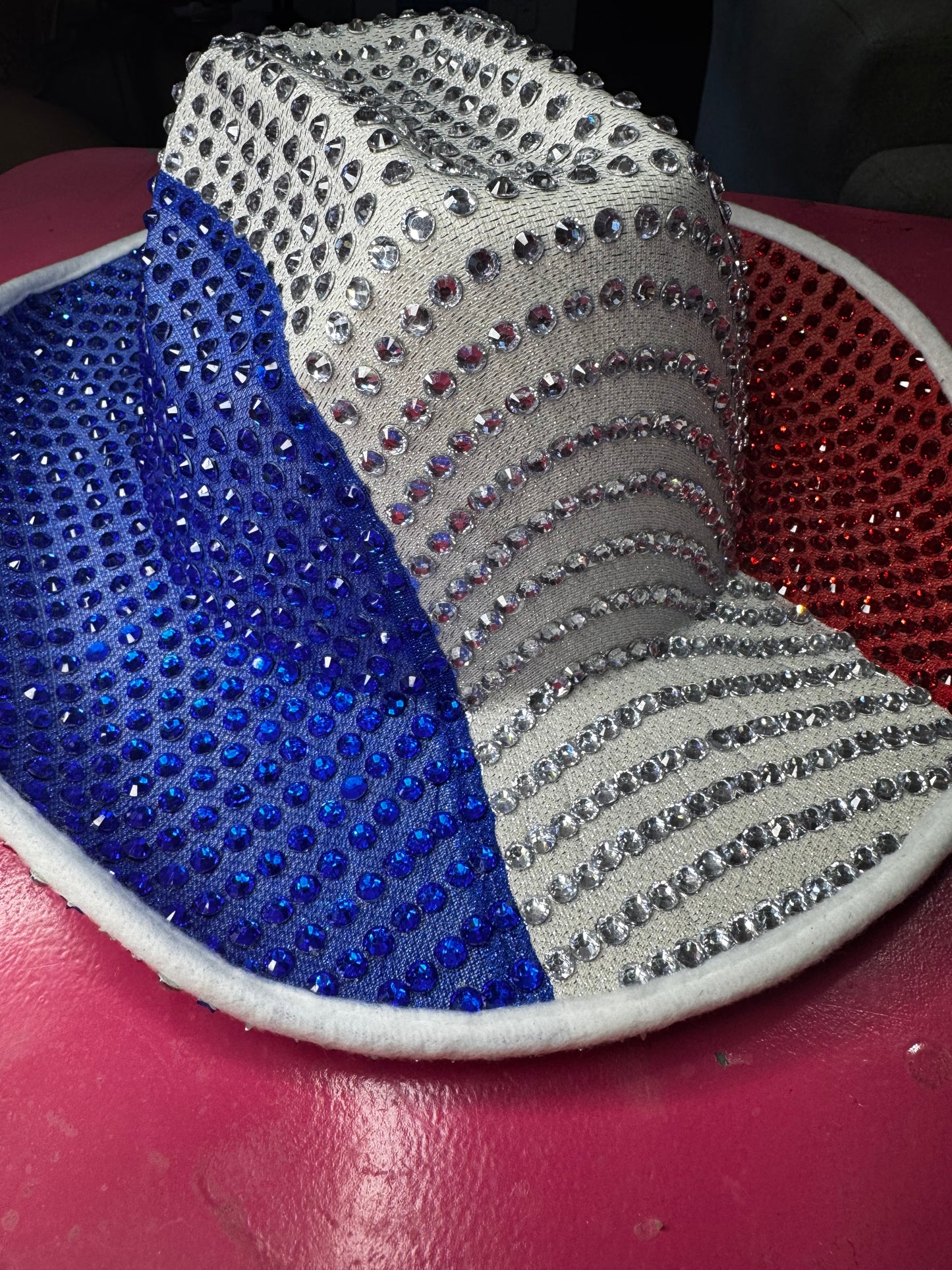 The Viva La Bombshell Burlesque Rhinestoned Cowboy Hat features silver, blue, and red rhinestones in three sections, sparkling brilliantly for a vibrant and eye-catching look. It is shown resting on a pink surface.