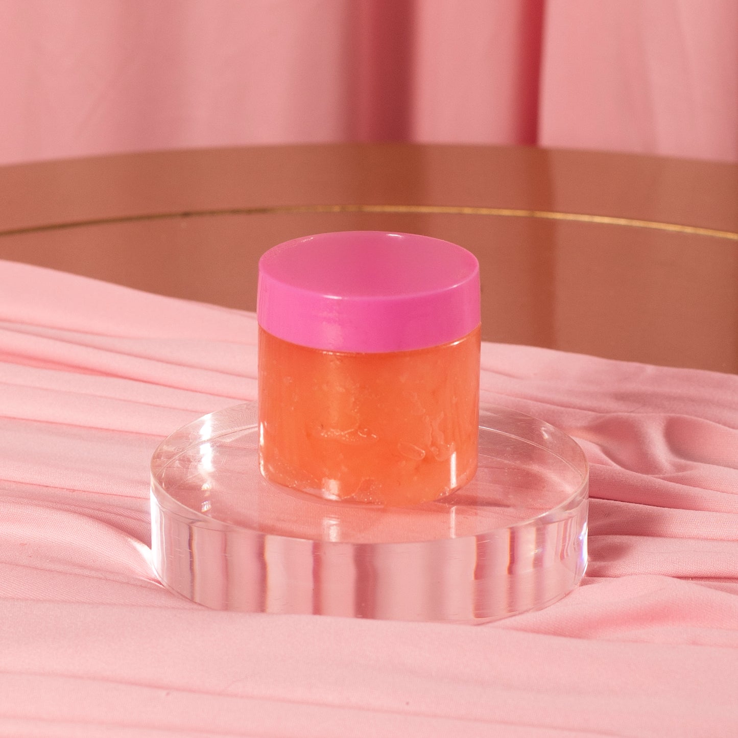 The Diamond Set by Viva La Bombshell Burlesque features a small jar of body scrub with a bright pink lid and orange formula, displayed on a clear round platform amid soft pink fabric and a rose gold backdrop.