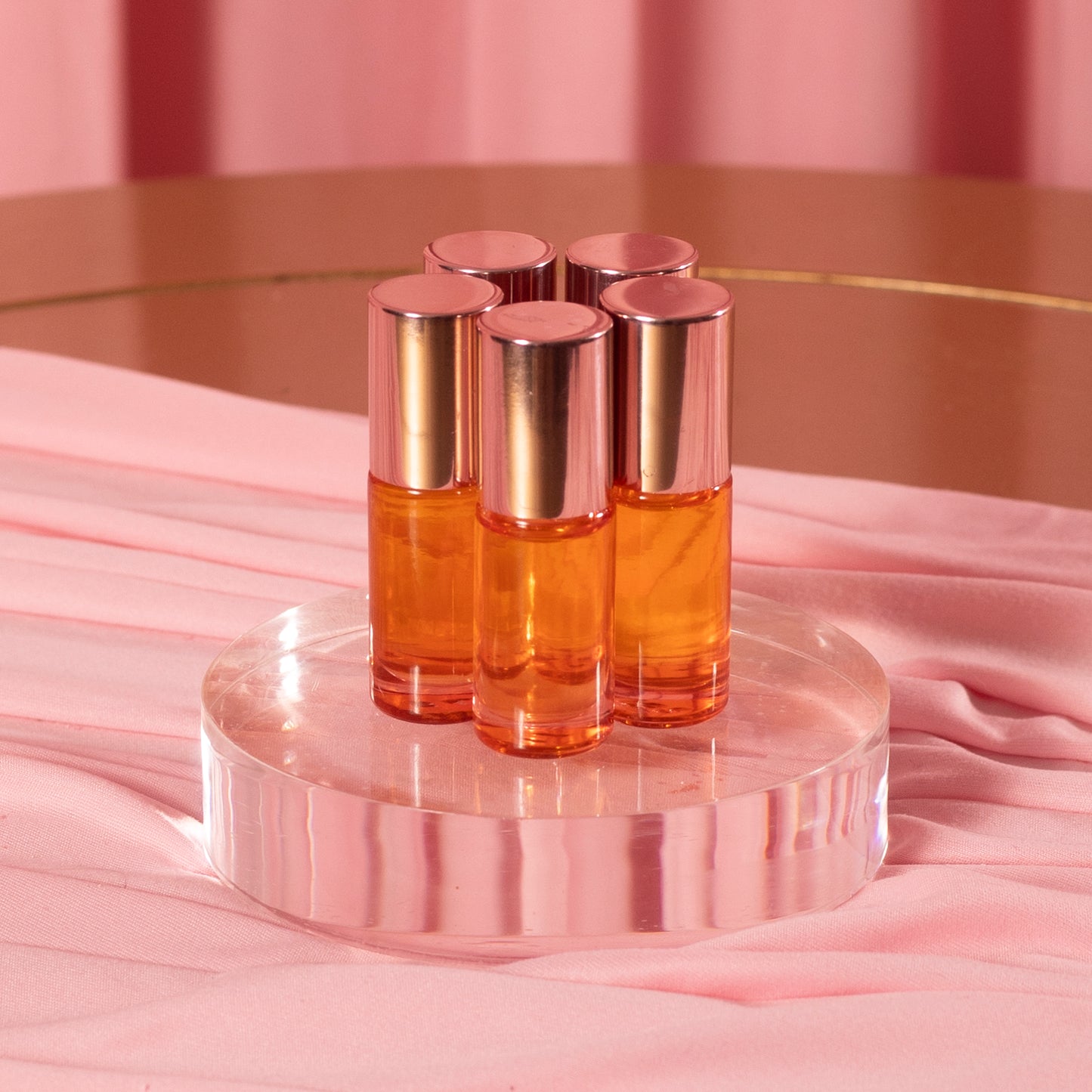 Five Viva La Bombshell Burlesque Cuticle Oil bottles, each with amber oil and rose gold caps, are arranged on a clear circular platform against a soft pink fabric backdrop.