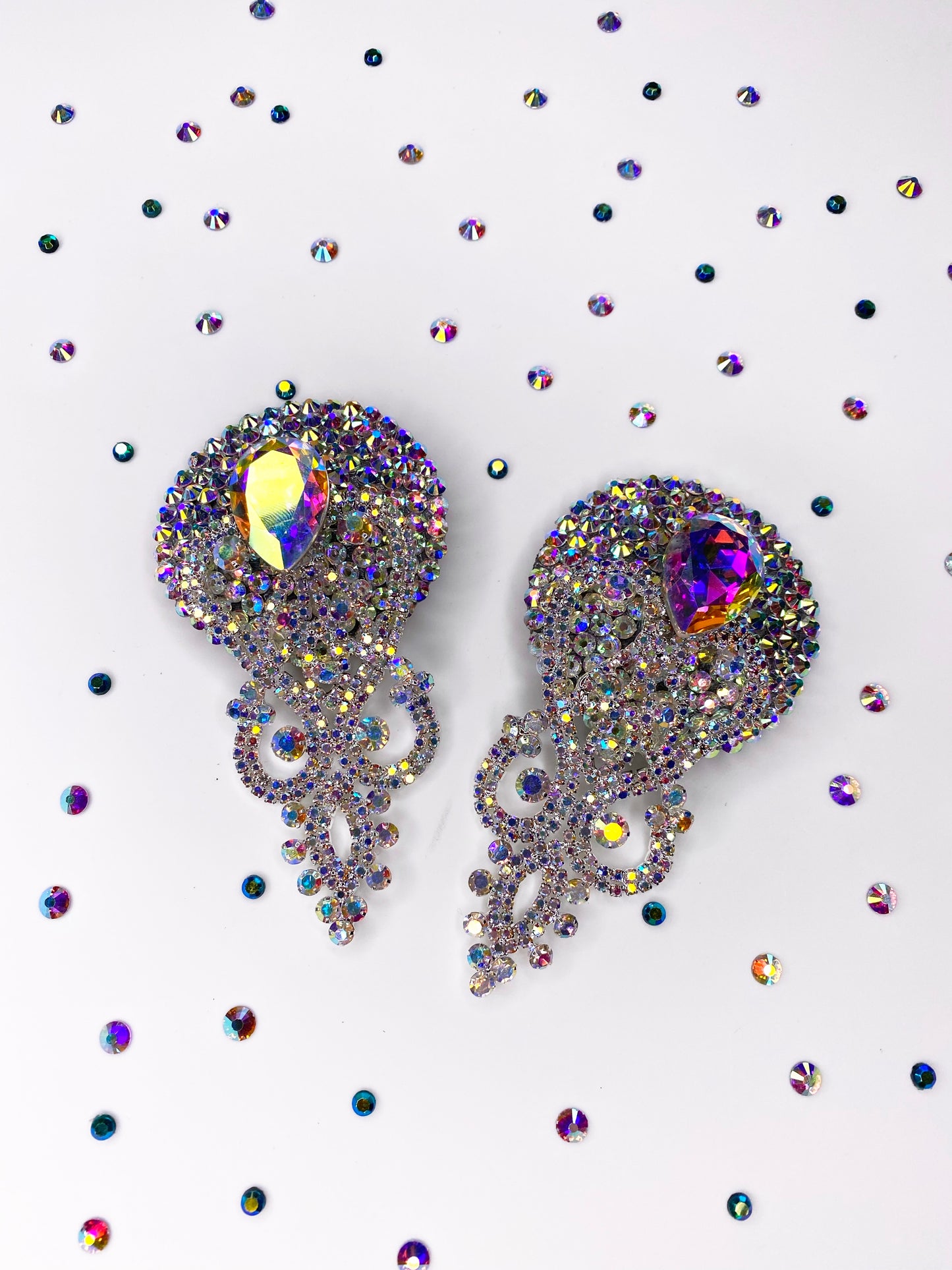Two ornate Showgirl Pasties by Viva La Bombshell Burlesque, featuring iridescent jewels and intricate designs, are displayed on a white surface surrounded by scattered crystal rhinestones of varying sizes.
