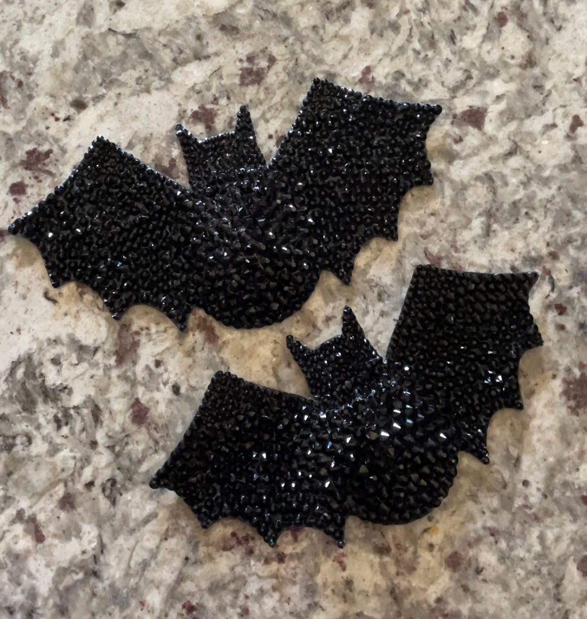Two black, bat-shaped Batty Pasties from Viva La Bombshell Burlesque, adorned with shiny rhinestones, rest on a speckled surface—ideal for adding sparkle to your spooky pasties collection.