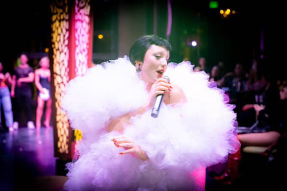 A performer wows the audience during an electrifying drag show, wearing Viva La Bombshell Burlesque’s The De'Vil Tulle Boa and a dramatic white costume, singing into a microphone on a colorful, dimly lit stage.