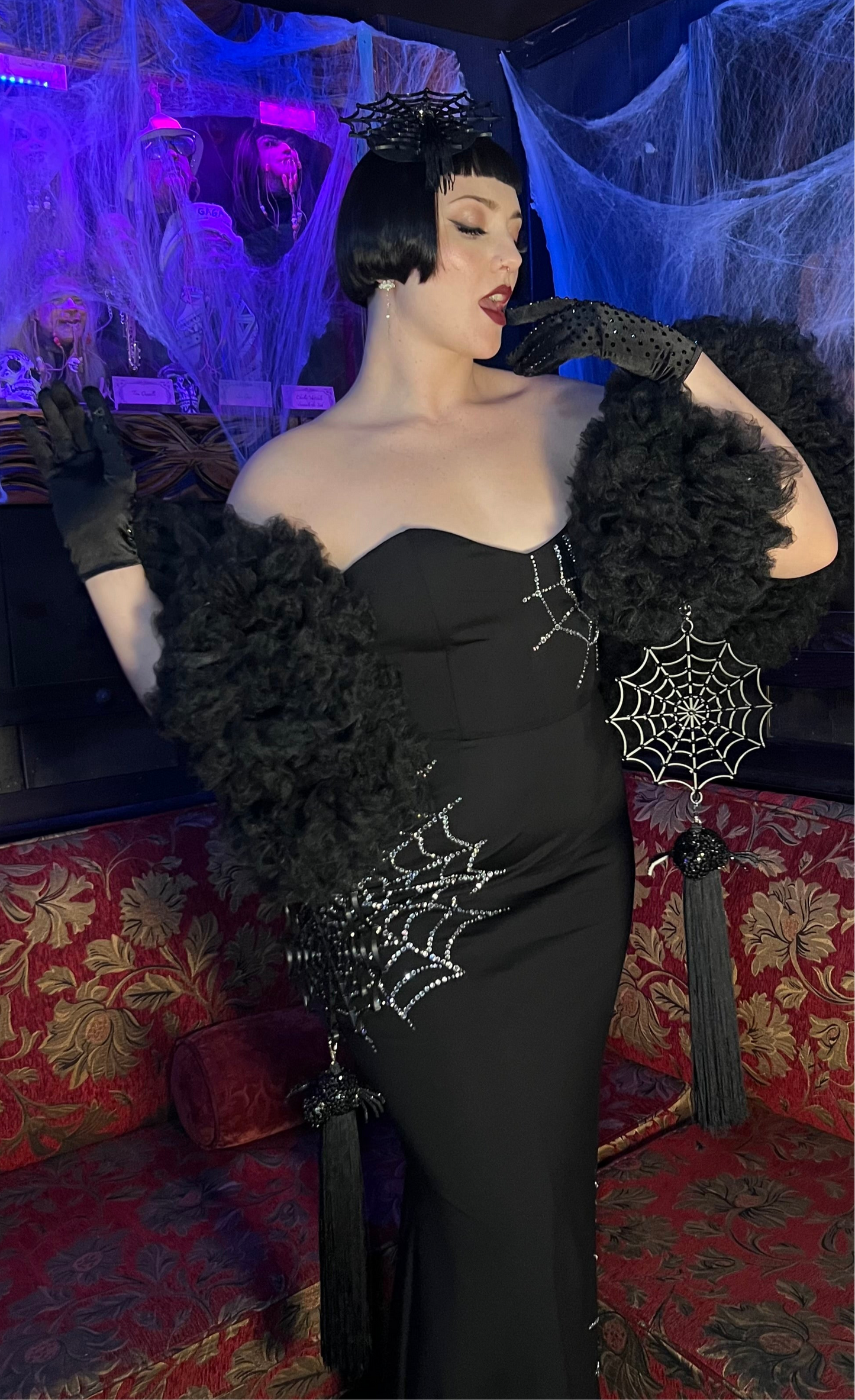 Wearing the Spider Boa by Viva La Bombshell Burlesque, a person poses glamorously on a red couch in a black rhinestone web gown with gloves and a headpiece, surrounded by Halloween cobwebs for spooky season.