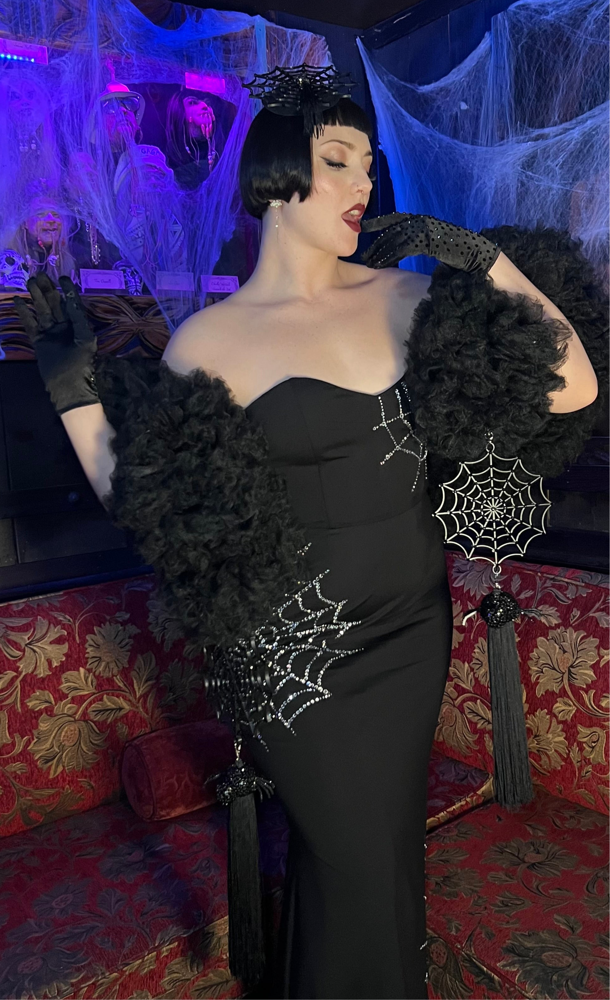 Wearing the Spider Boa by Viva La Bombshell Burlesque, a person poses glamorously on a red couch in a black rhinestone web gown with gloves and a headpiece, surrounded by Halloween cobwebs for spooky season.