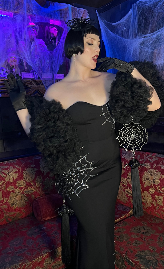 Wearing the Spider Boa by Viva La Bombshell Burlesque, a person poses glamorously on a red couch in a black rhinestone web gown with gloves and a headpiece, surrounded by Halloween cobwebs for spooky season.