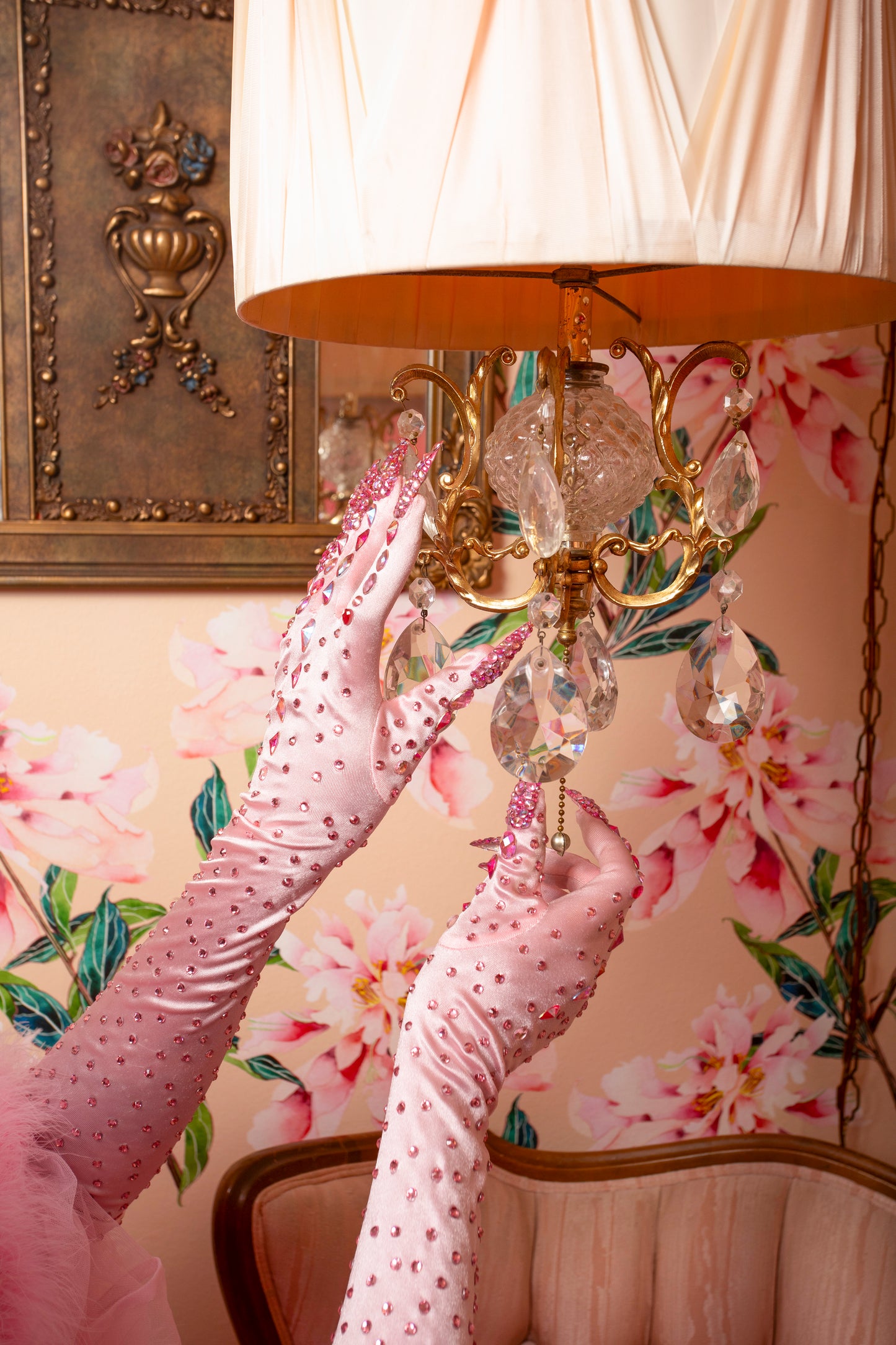 Wearing Viva La Bombshell Burlesque’s Kitty Claw Gloves (AB Rhinestones) - Original, a person adjusts a gold lamp with a pleated shade. The floral wallpaper and vintage chair in the background evoke retro elegance.
