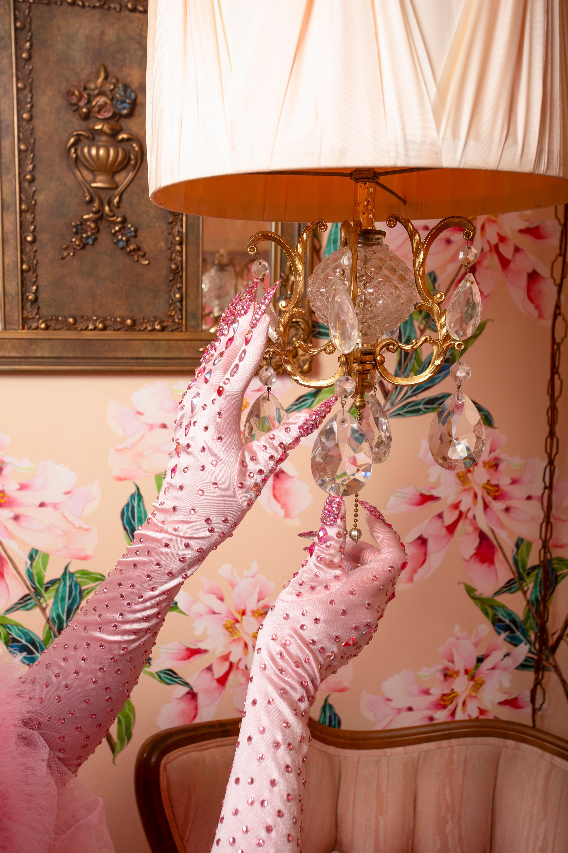 Wearing Viva La Bombshell Burlesque’s Kitty Claw Gloves (AB Rhinestones) - Original, a person adjusts a gold lamp with a pleated shade. The floral wallpaper and vintage chair in the background evoke retro elegance.