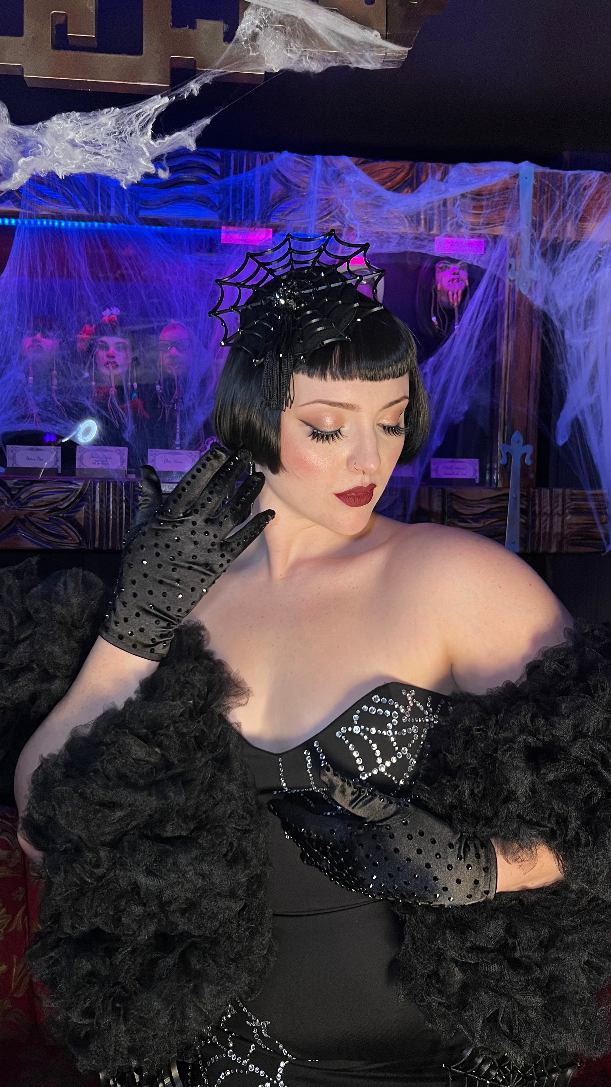 A woman in glamorous vintage style poses indoors, wearing a black dress, sparkling details, polka-dot gloves, a dramatic fur stole, and the Arachné Noir Fascinator by Viva La Bombshell Burlesque amid Halloween decorations.