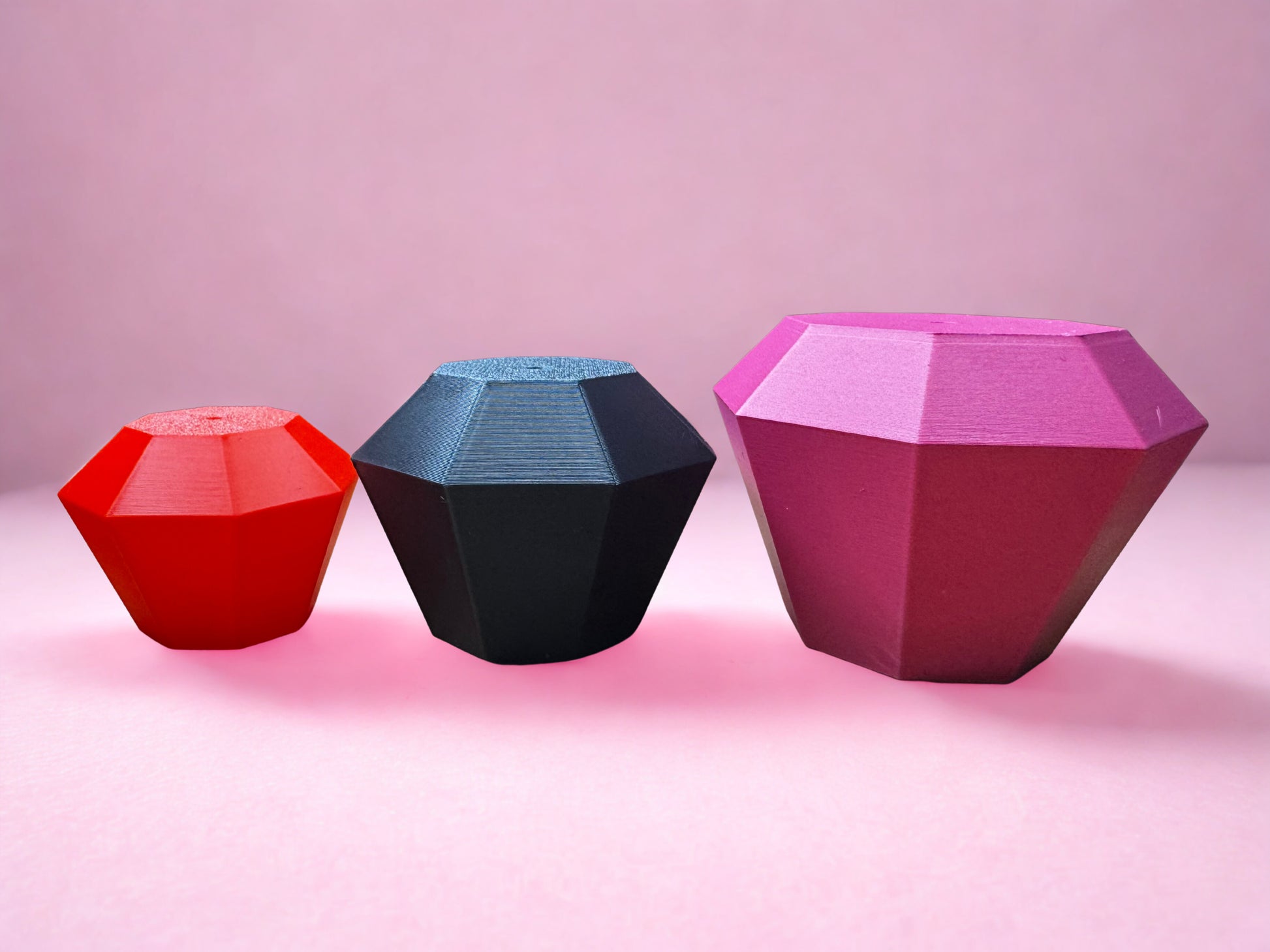 Three faceted objects in red, black, and pink are arranged on a pink surface, echoing the dazzling design of Tassel Toppers - Boa by Viva La Bombshell Burlesque—perfect inspiration for creative party decor.