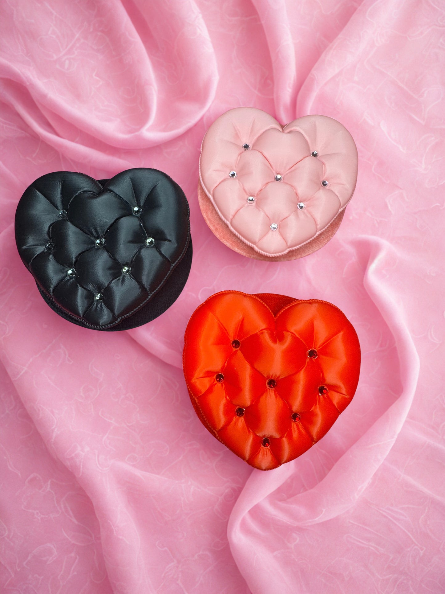 Three heart-shaped Tufted Heart Fascinator boxes by Viva La Bombshell Burlesque—black, pink, and red—rest on a soft pink textured fabric background, echoing the handmade look of a unique burlesque accessory.
