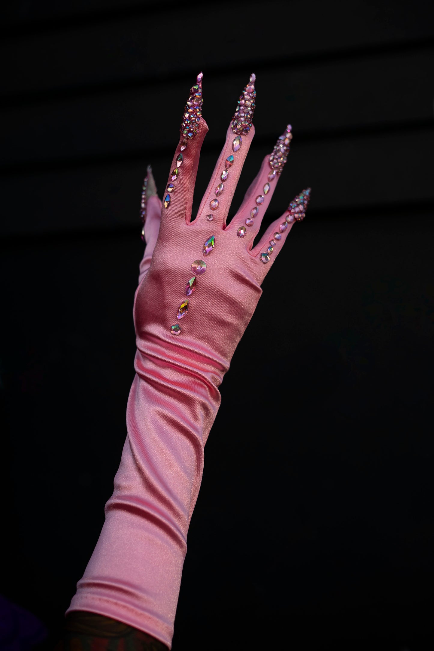 A hand with rhinestone-encrusted stiletto nails elegantly models Viva La Bombshell Burlesque’s Cash Claw Gloves—long pink gloves adorned with sparkling rhinestones.