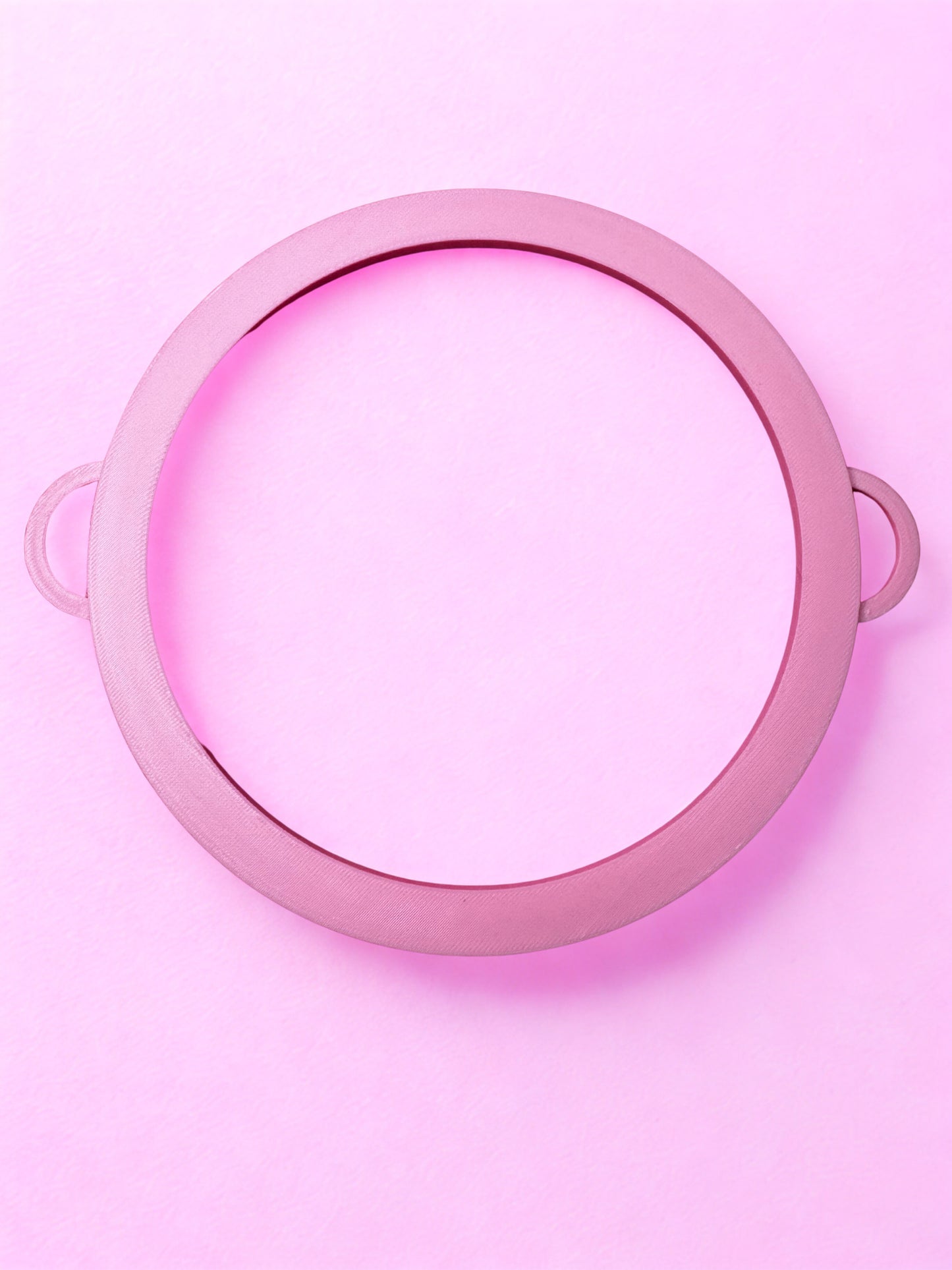 The Circle Bangle - Boa Attachment by Viva La Bombshell Burlesque is a pink circular frame with two handles, placed on a pastel pink background. The center is empty, showcasing its modern bangle-inspired design.