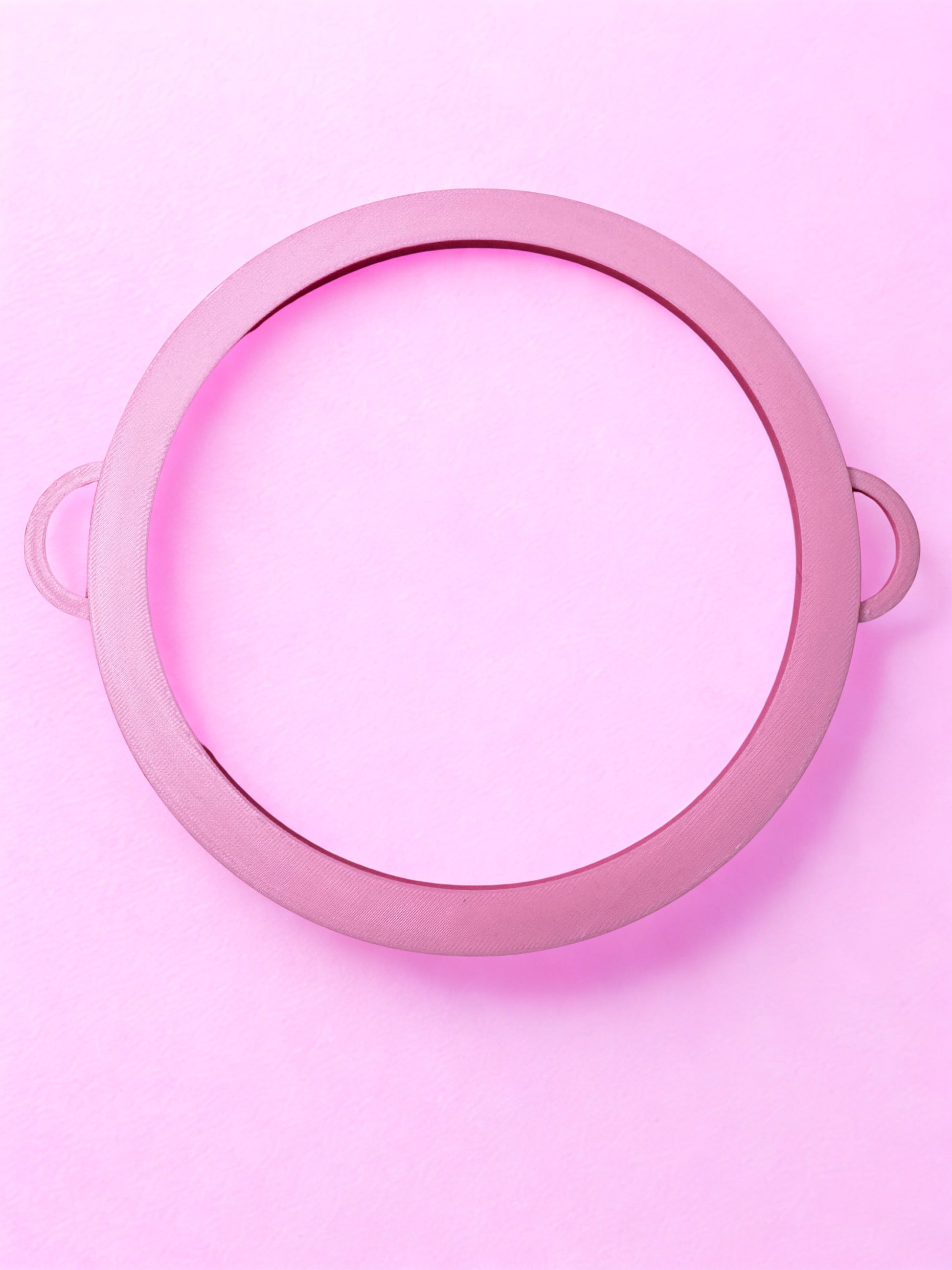 The Circle Bangle - Boa Attachment by Viva La Bombshell Burlesque is a pink circular frame with two handles, placed on a pastel pink background. The center is empty, showcasing its modern bangle-inspired design.