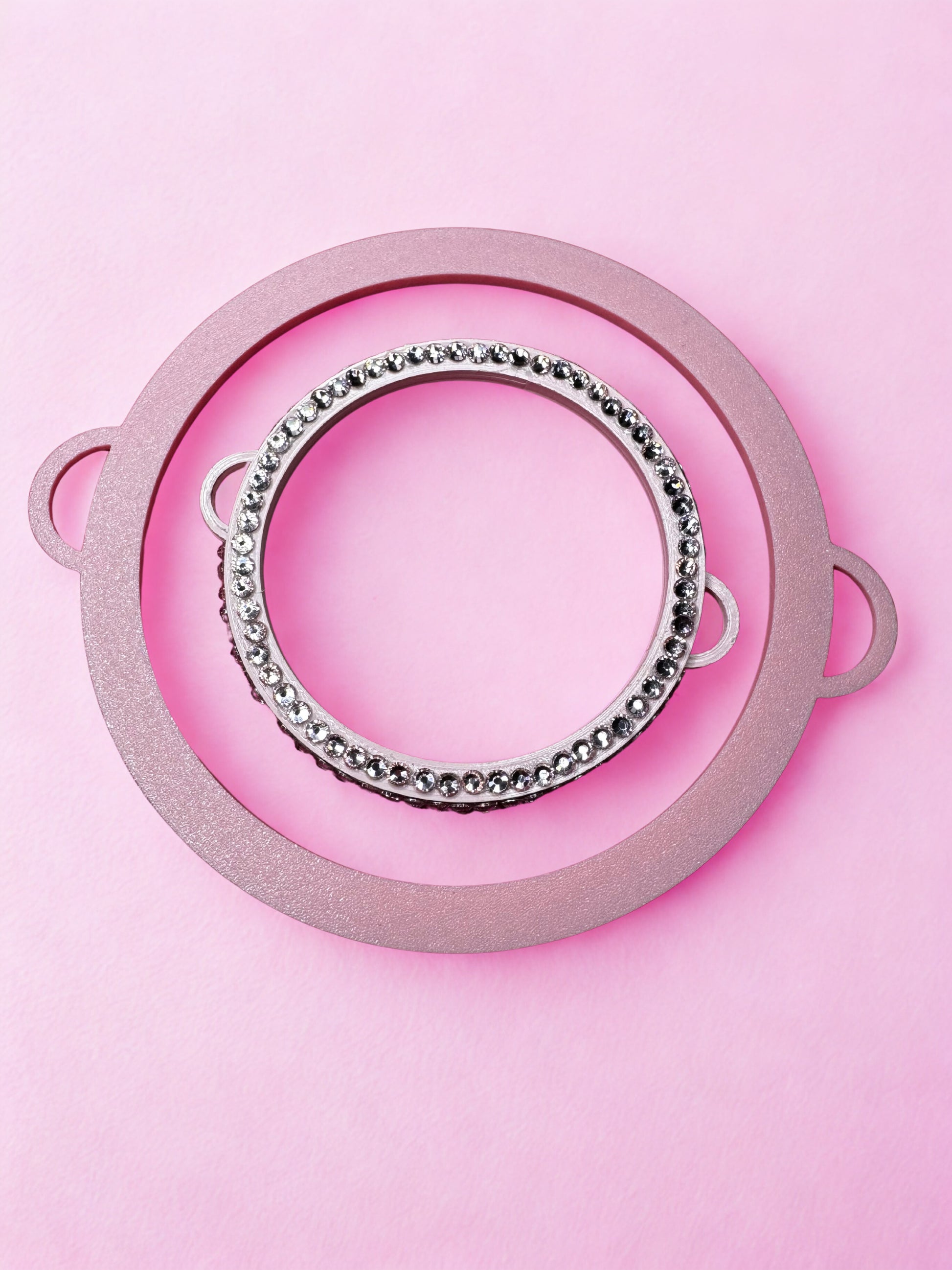 The Viva La Bombshell Burlesque Circle Bangle - Boa Attachment features a round pink frame with handles, a metallic ring adorned with shiny silver beads, and makes the ideal display for statement jewelry on a pastel pink background.