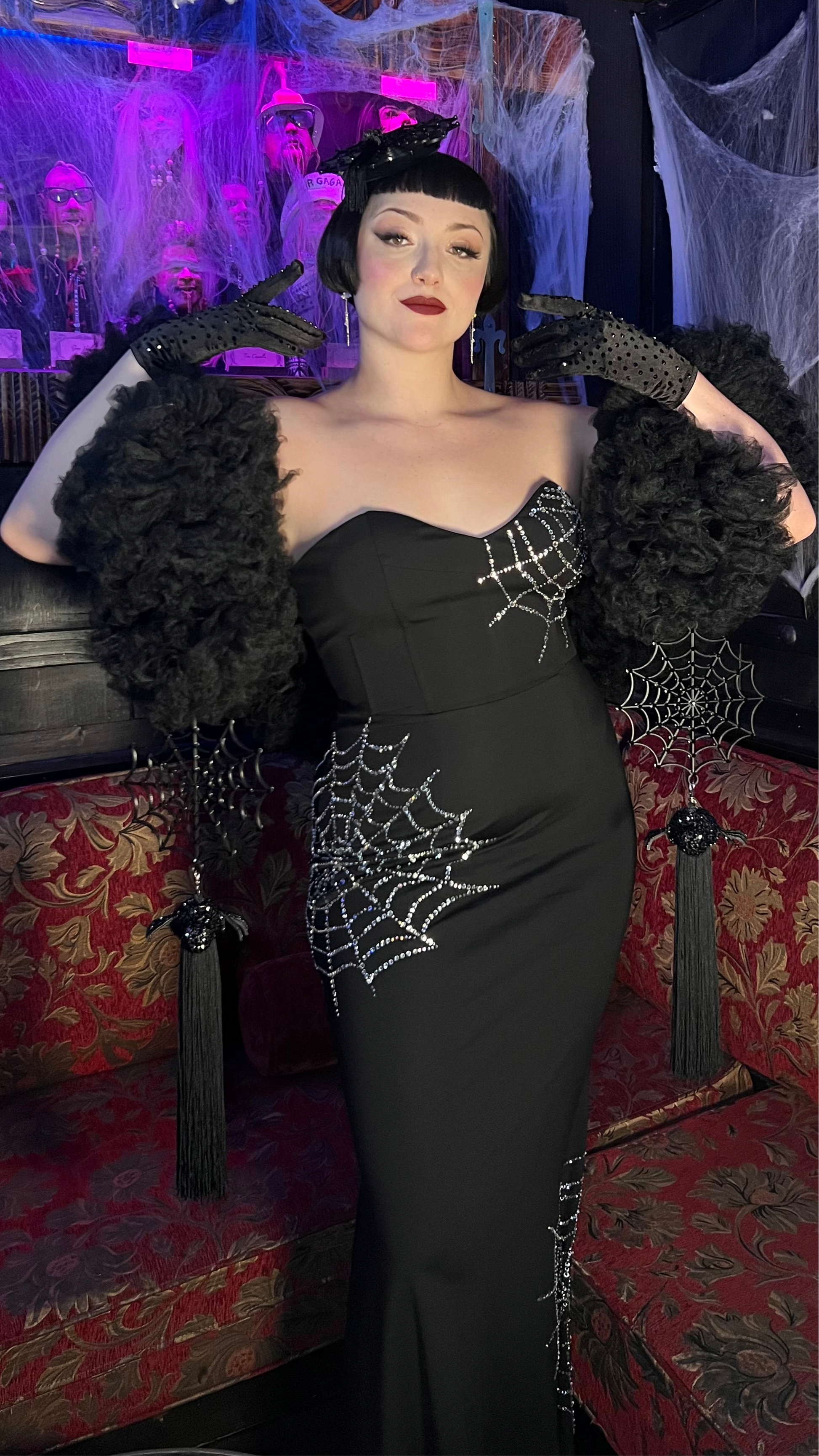 A person poses on a red couch wearing Viva La Bombshell Burlesque's Spider Boa—a strapless black gown with rhinestone web details and dramatic feathered sleeves, short black hair, dark makeup, and cobweb decor—perfect for spooky season.
