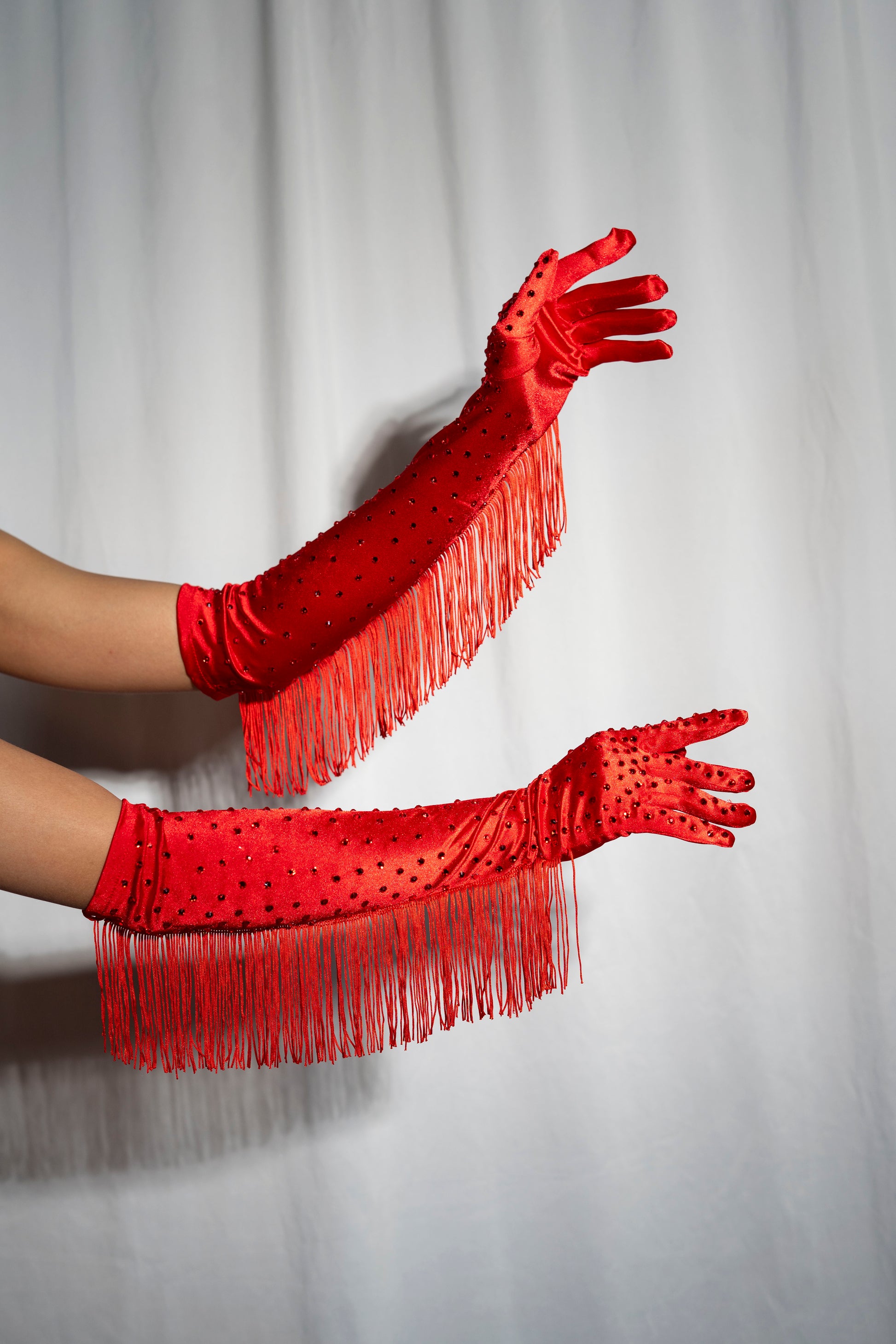 A pair of Viva La Bombshell Burlesque Fringe Gloves in red satin, detailed with fringe and small black beads, are elegantly displayed with arms extended against a plain white curtain backdrop.