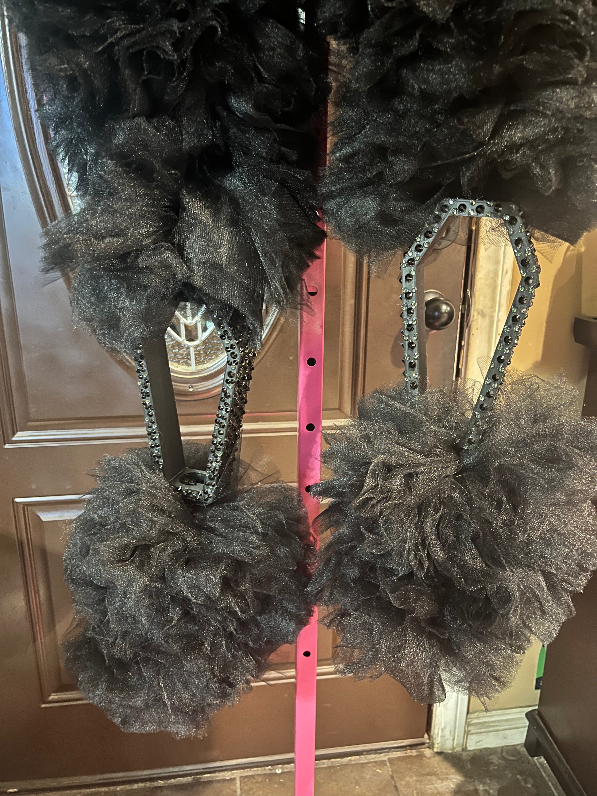 Close-up of two black tulle pom-poms attached to black rhinestone coffin bangles, hanging from a bright pink vertical pole, with a brown door in the background.
