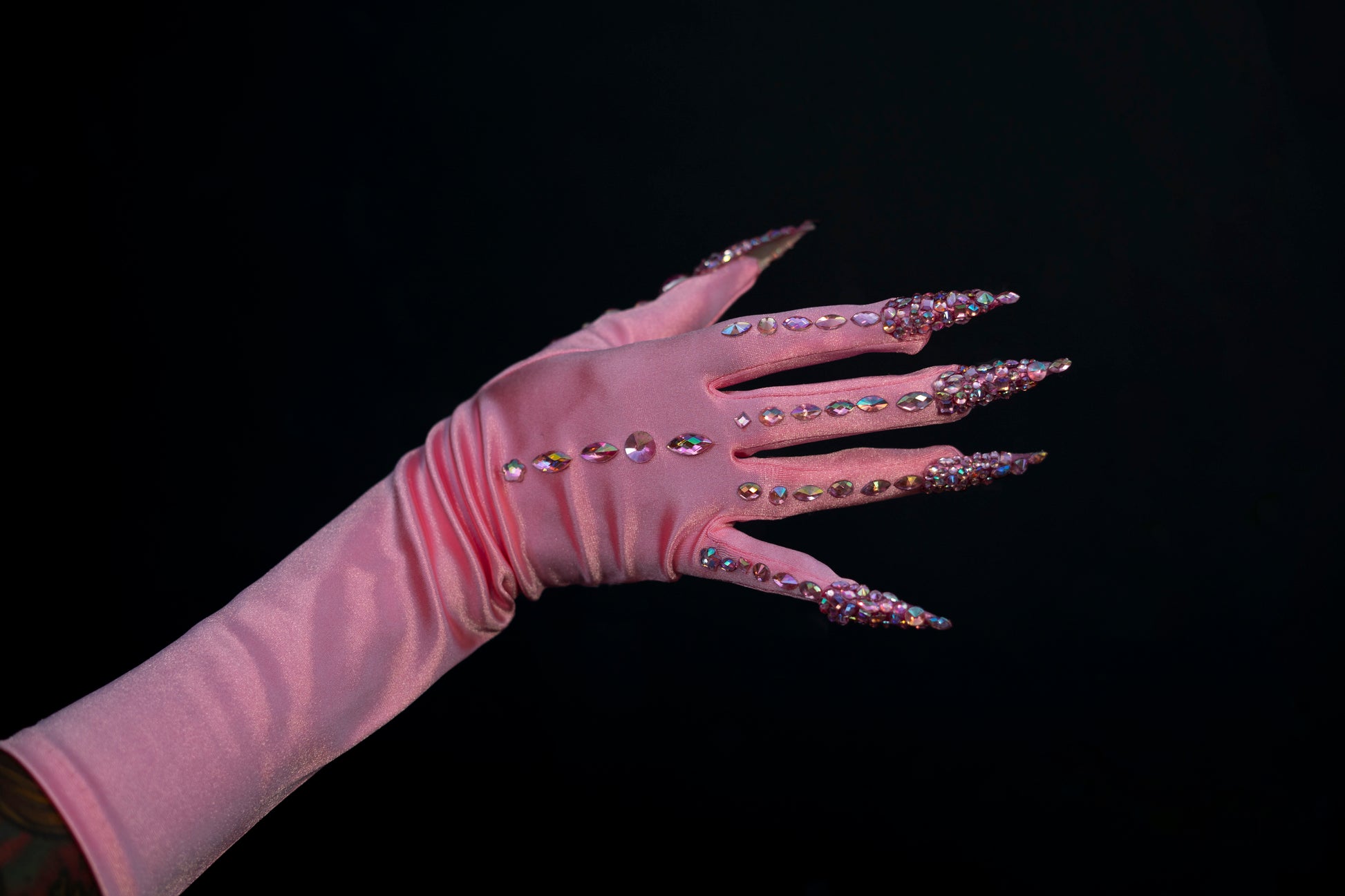 A hand in Viva La Bombshell Burlesque’s Cash Claw Gloves—shiny pink with rhinestone stiletto nails and sparkling stones—stands out against a solid black background.