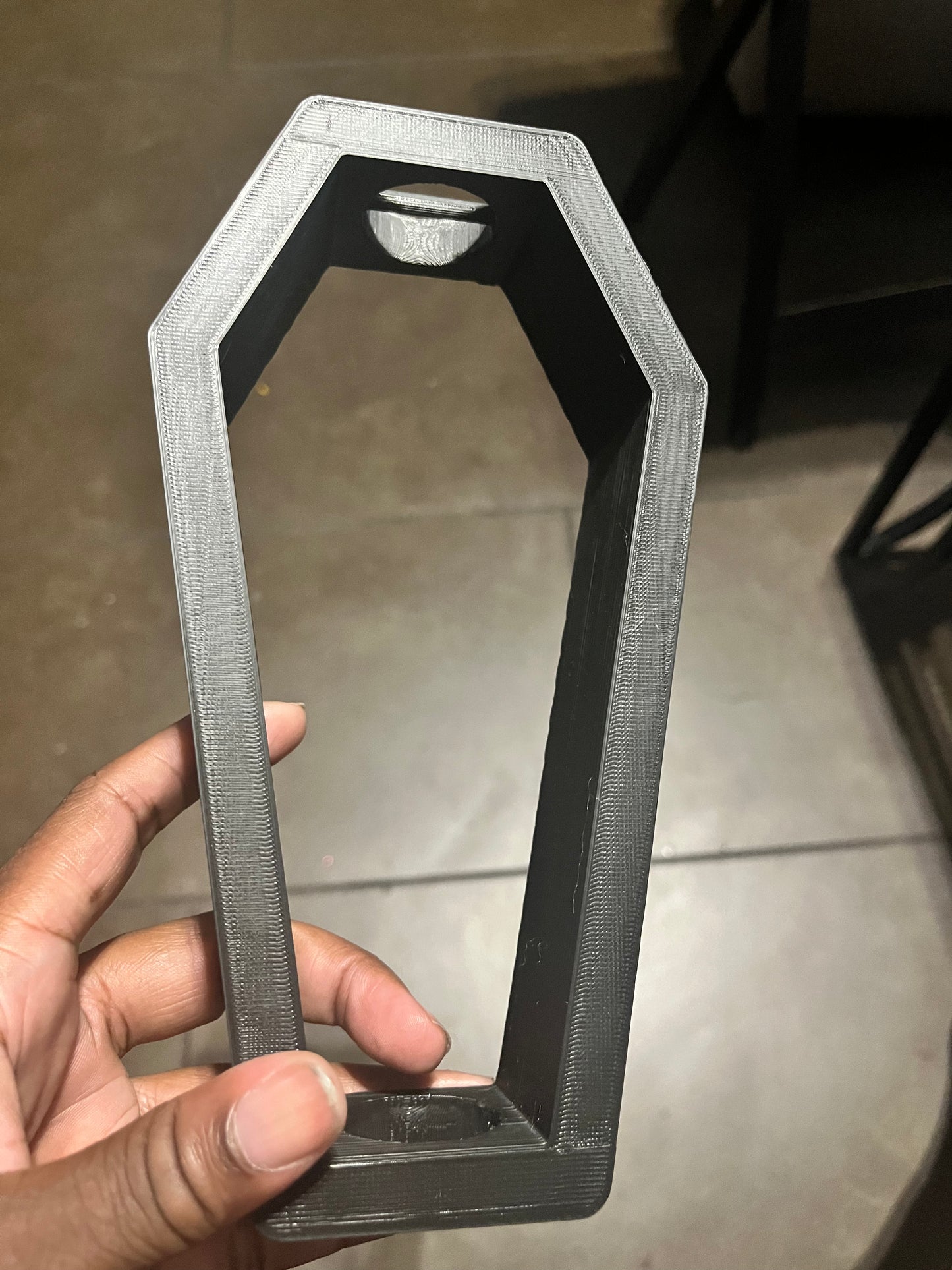 A hand holds a black, 3D-printed bangle shaped like a coffin with an open center. The object has straight edges and a smooth surface, and is being held indoors over a tiled floor.