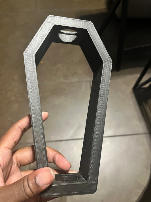 A hand holds a black, 3D-printed bangle shaped like a coffin with an open center. The object has straight edges and a smooth surface, and is being held indoors over a tiled floor.