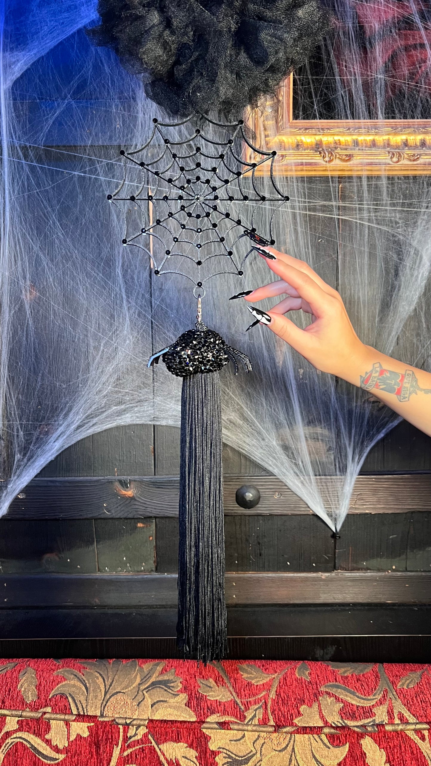 A hand with long black nails holds the Viva La Bombshell Burlesque Spider Boa Attachment, a black beaded spider tassel fringe with web design, radiating spooky glamour against a dark background with rhinestone web appliqués and a gold frame.