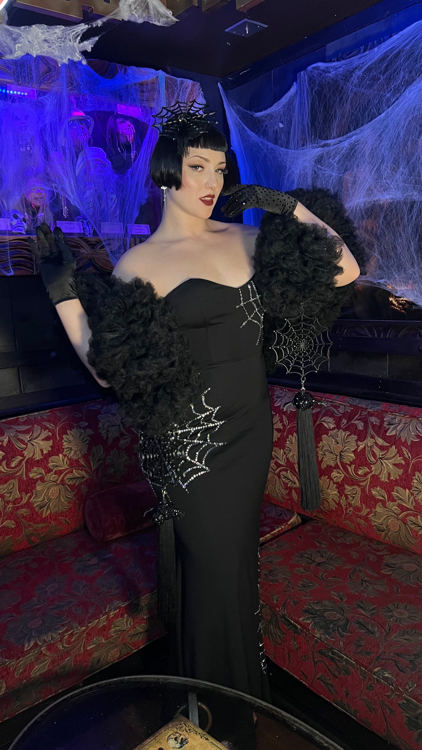 A person in a glamorous black gown with rhinestone webbing poses in a spooky-themed lounge. Wearing a short black wig, dark lipstick, gloves, and Viva La Bombshell Burlesque’s Spider Boa draped dramatically over their shoulders.