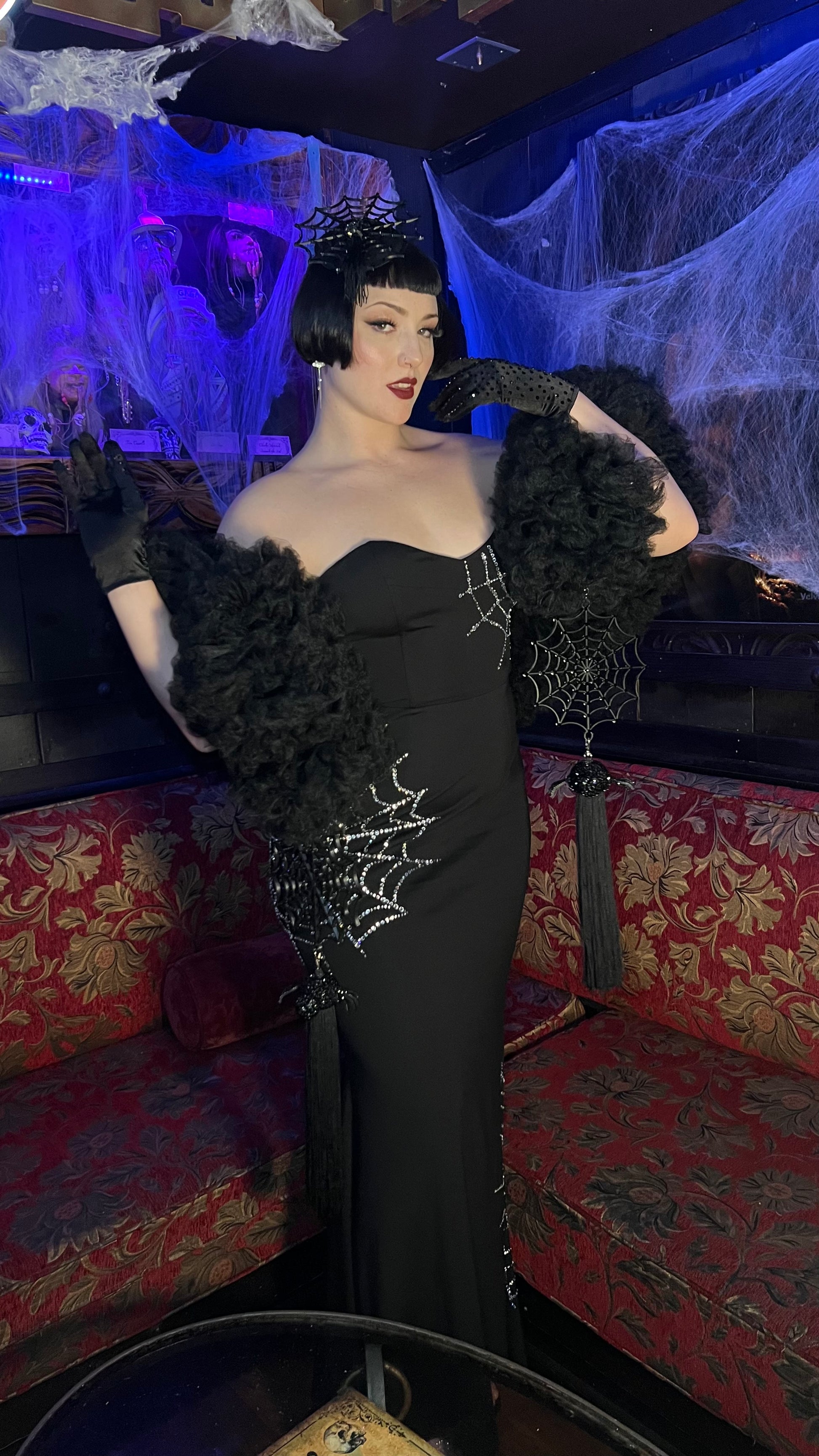 A person in a glamorous black gown with rhinestone webbing poses in a spooky-themed lounge. Wearing a short black wig, dark lipstick, gloves, and Viva La Bombshell Burlesque’s Spider Boa draped dramatically over their shoulders.