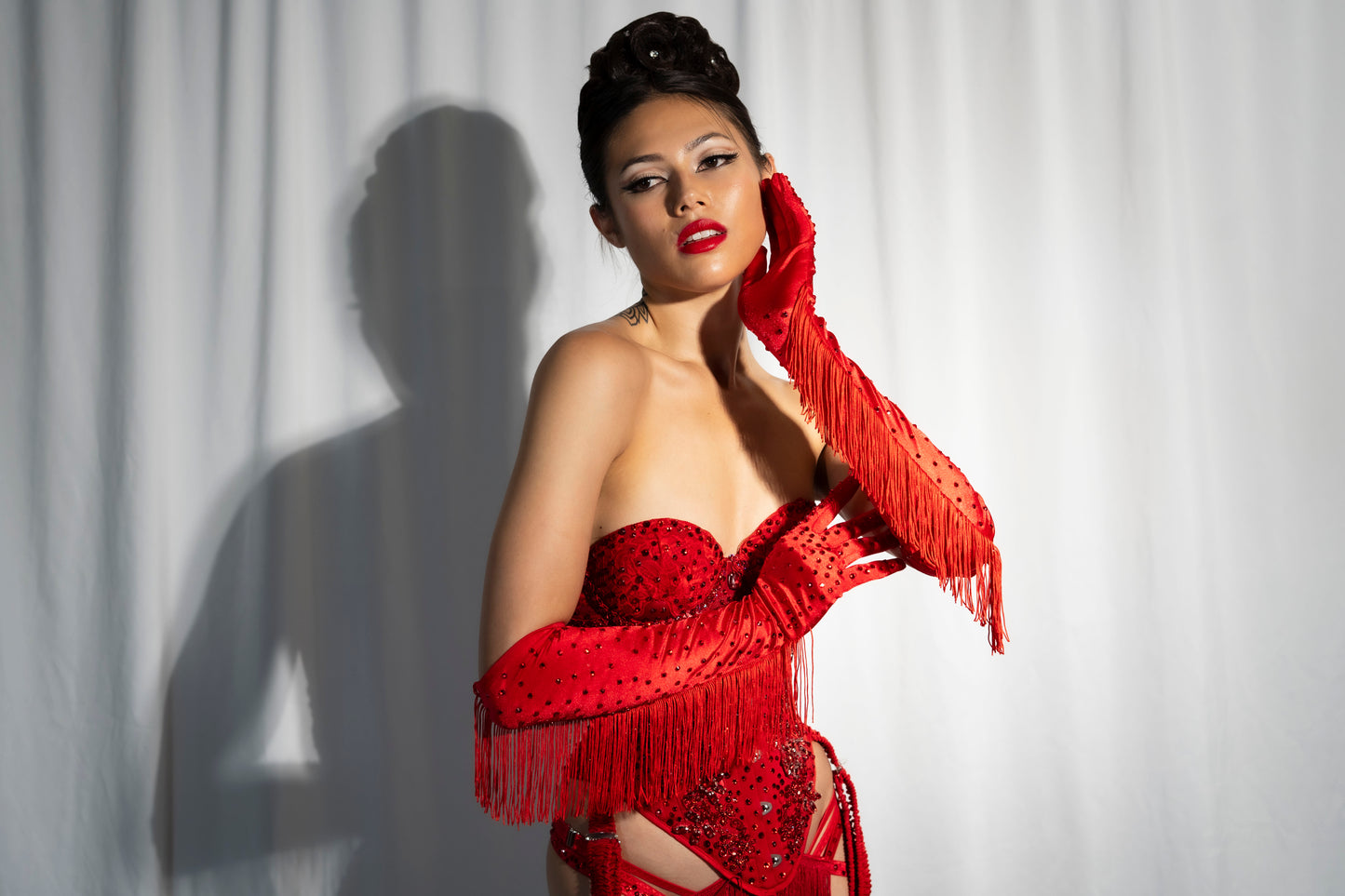 A woman poses before a white curtain in Viva La Bombshell Burlesque’s Fringe Gloves—elbow-length, rhinestoned, and perfect for stage performance—her elegant updo and bold makeup highlighted as she touches her face, casting a dramatic shadow.