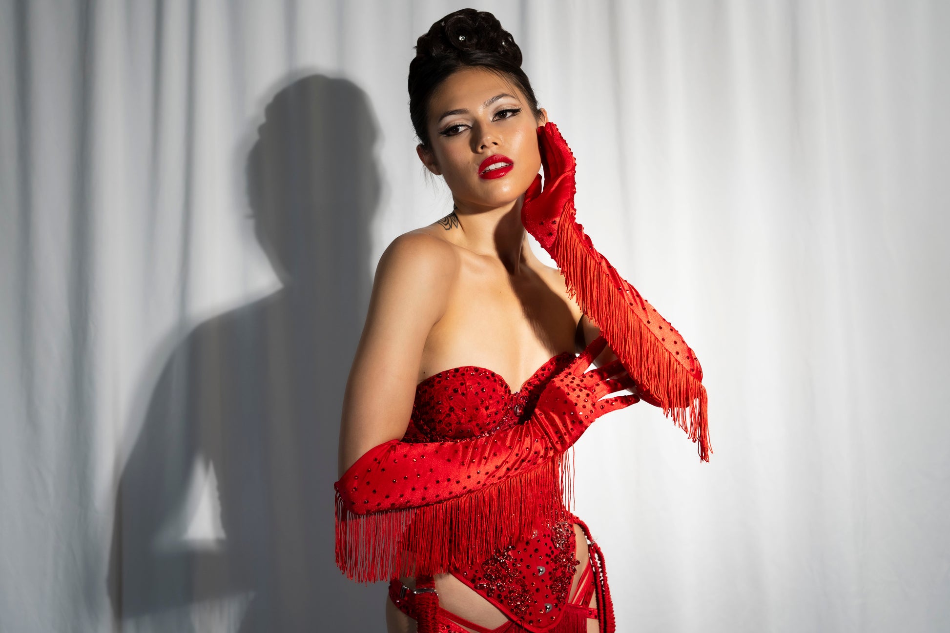 A woman poses before a white curtain in Viva La Bombshell Burlesque’s Fringe Gloves—elbow-length, rhinestoned, and perfect for stage performance—her elegant updo and bold makeup highlighted as she touches her face, casting a dramatic shadow.