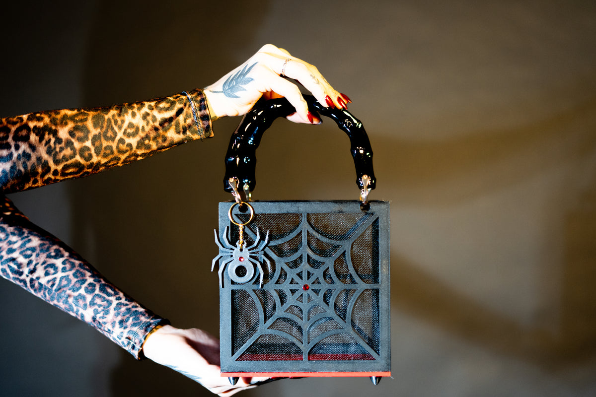 A tattooed hand with red nails holds The Widow – Spiderweb Purse by Viva La Bombshell Burlesque, a square handbag adorned with a striking spiderweb design, a spider charm, and elegant bamboo handles; the person wears a leopard-print sleeve.