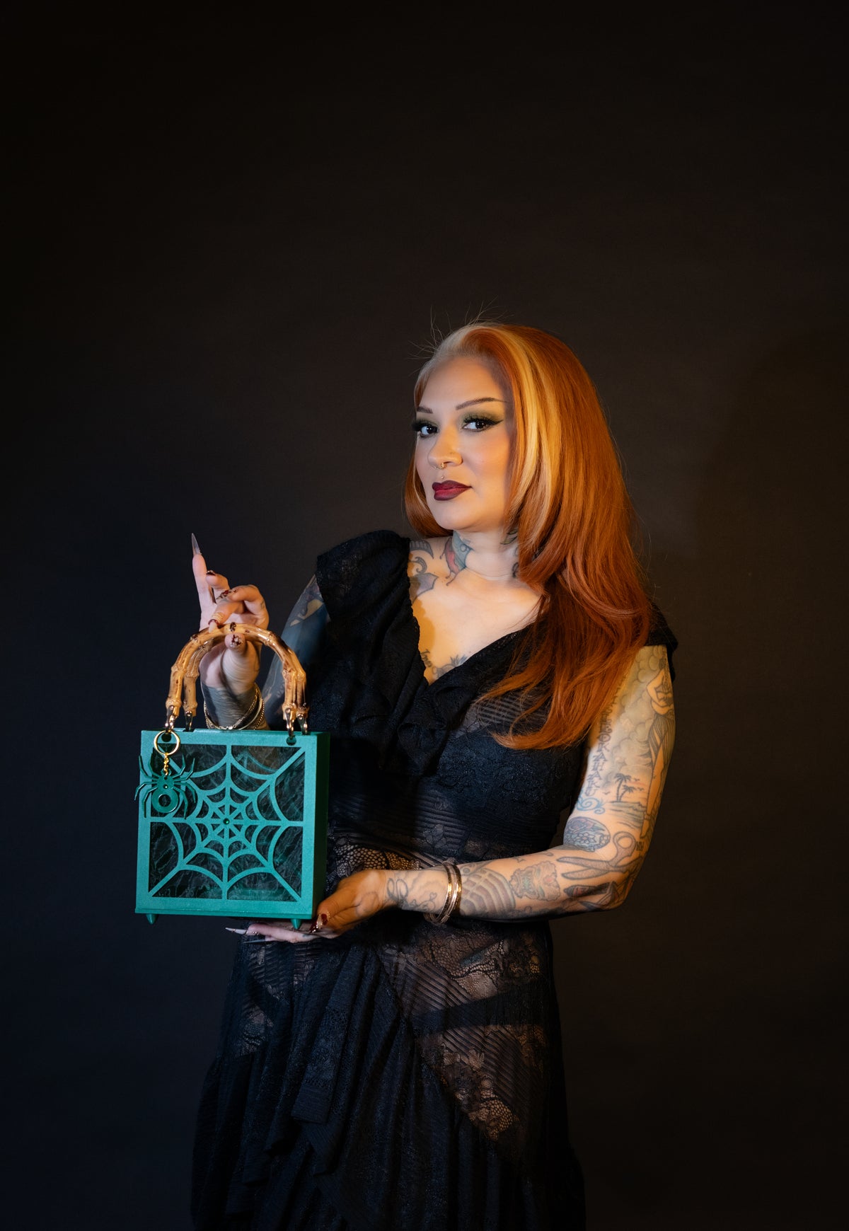 A woman with long red hair and tattoos wears a black lace dress and holds The Widow – Spiderweb Purse by Viva La Bombshell Burlesque, a green handbag with bamboo handles and a striking spiderweb design, against a dark background.