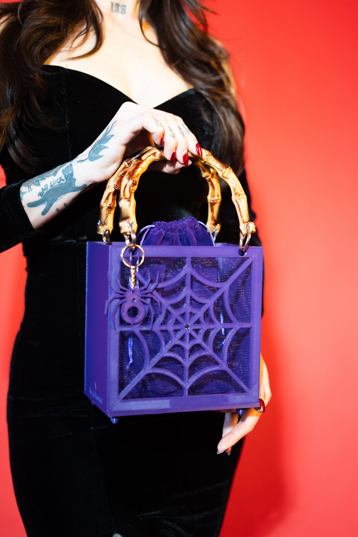 A person in a black dress holds The Widow – Spiderweb Purse by Viva La Bombshell Burlesque, showcasing its bold purple 3D-printed spiderweb design and chic bamboo handles, while standing against a vibrant red background.