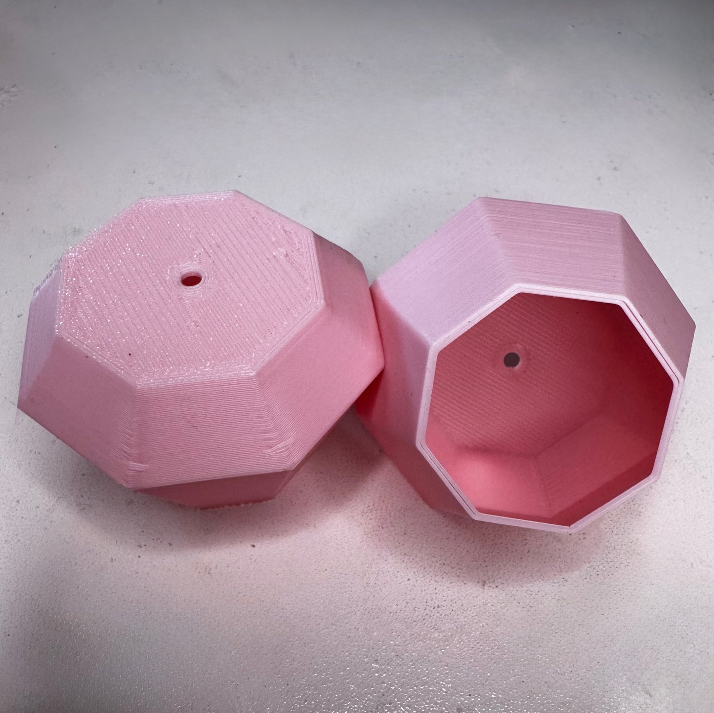 Two pink, geometric, 3D-printed plant pots with faceted sides and drainage holes from Viva La Bombshell Burlesque rest on a light surface—one upright, one on its side—perfect for displaying Tassel Toppers - Boa.