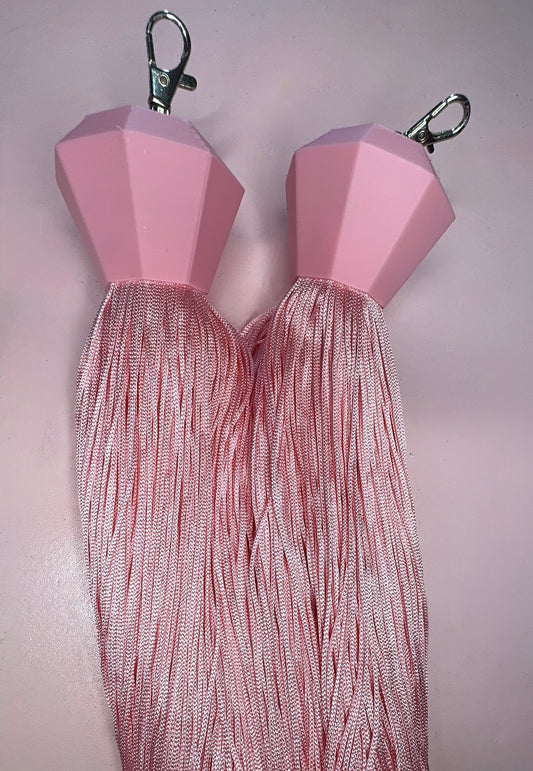 The Viva La Bombshell Burlesque Boa Tassels feature pink gem-shaped clip-ons with metal clasps attached to long, shiny pink tassels, displayed against a pale pink background.