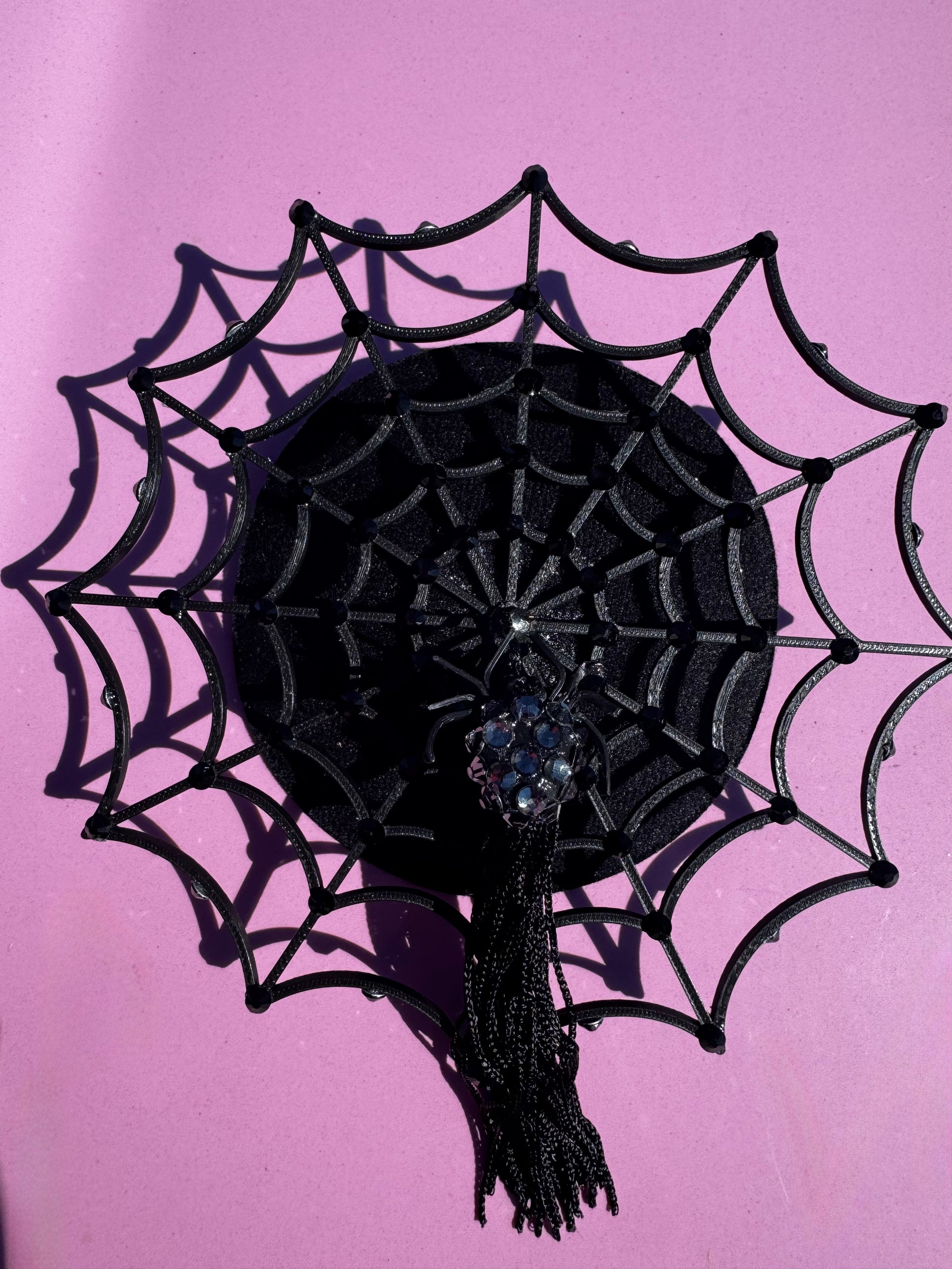 The Arachné Noir Fascinator by Viva La Bombshell Burlesque features a black spiderweb with a beaded spider center, shown on a bright pink background. This bold headpiece adds spooky flair to any costume or party look.