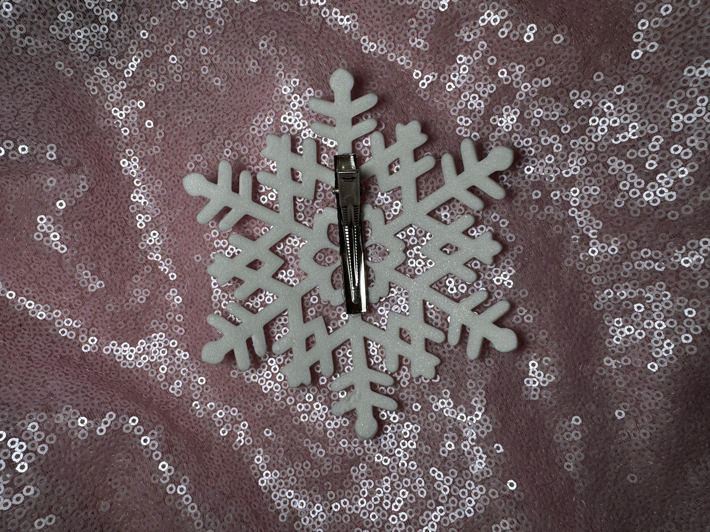 The Snow Queen Fascinator by Viva La Bombshell Burlesque is displayed on a white foam snowflake, resembling its unique style, set against a pink sequin fabric background.