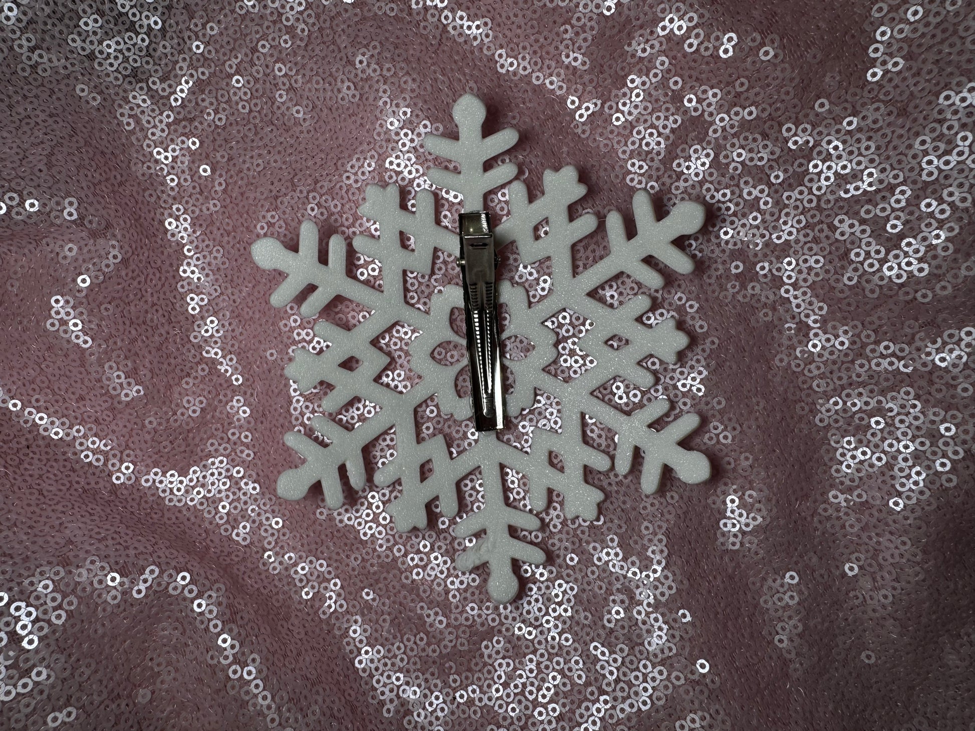 The Snow Queen Fascinator by Viva La Bombshell Burlesque is displayed on a white foam snowflake, resembling its unique style, set against a pink sequin fabric background.