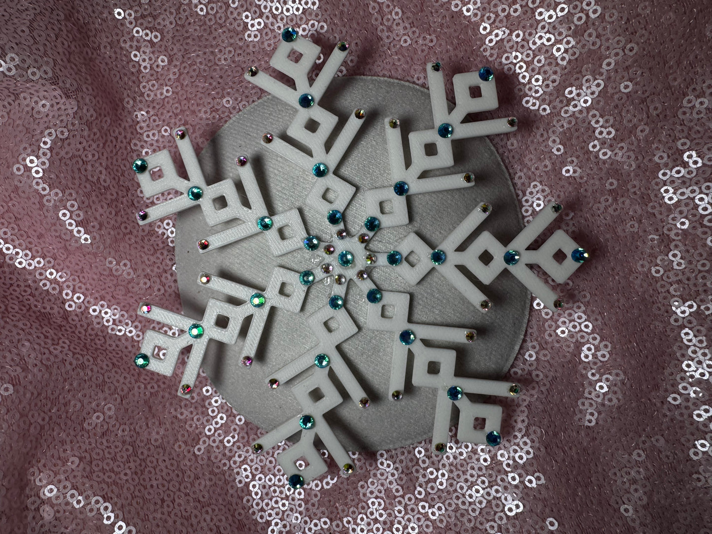 The Ice Queen Fascinator by Viva La Bombshell Burlesque, a white geometric snowflake adorned with colorful gems, is displayed on a pink sequin fabric background.