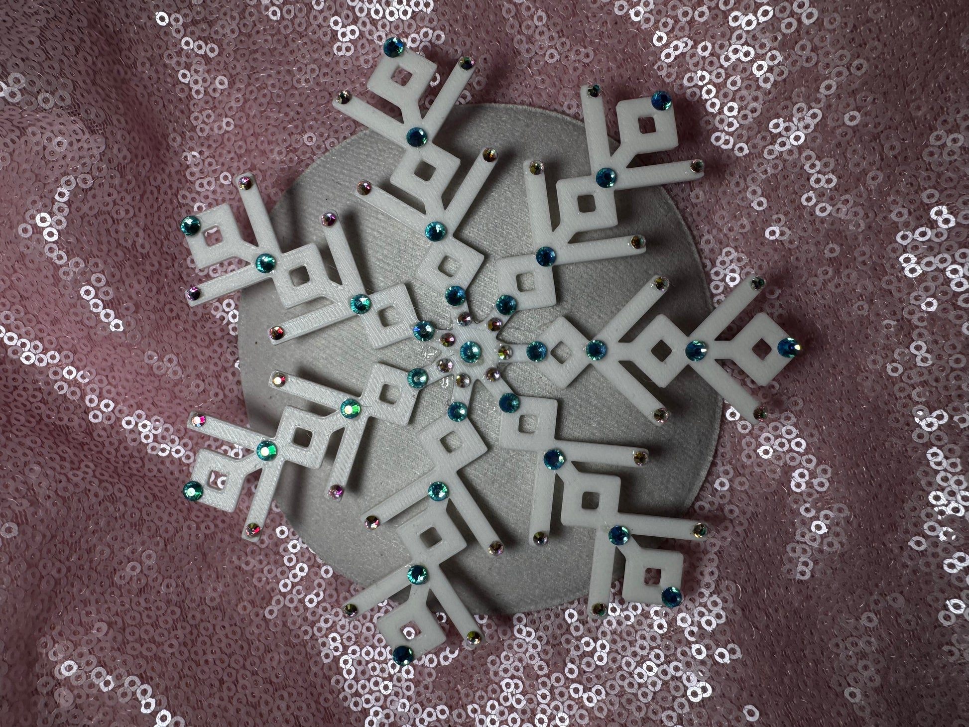 The Ice Queen Fascinator by Viva La Bombshell Burlesque, a white geometric snowflake adorned with colorful gems, is displayed on a pink sequin fabric background.