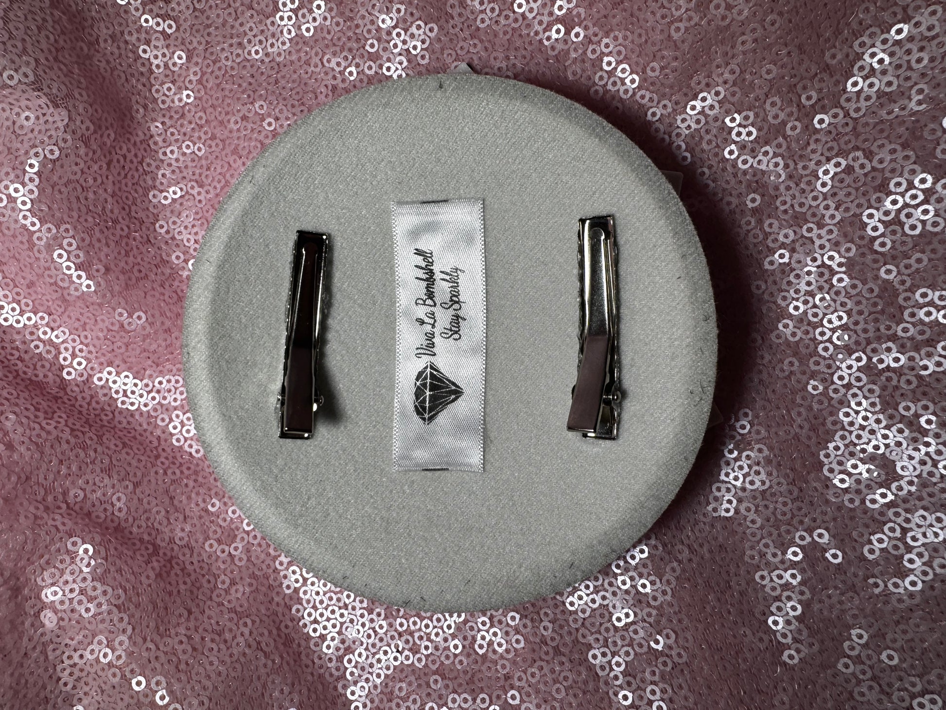 The Ice Queen Fascinator by Viva La Bombshell Burlesque is a light gray round snowflake piece with two silver hair clips, shown on pink sequin fabric. A white label reads "Miss Bella Bespoke, Stay Beautiful," featuring a diamond logo.