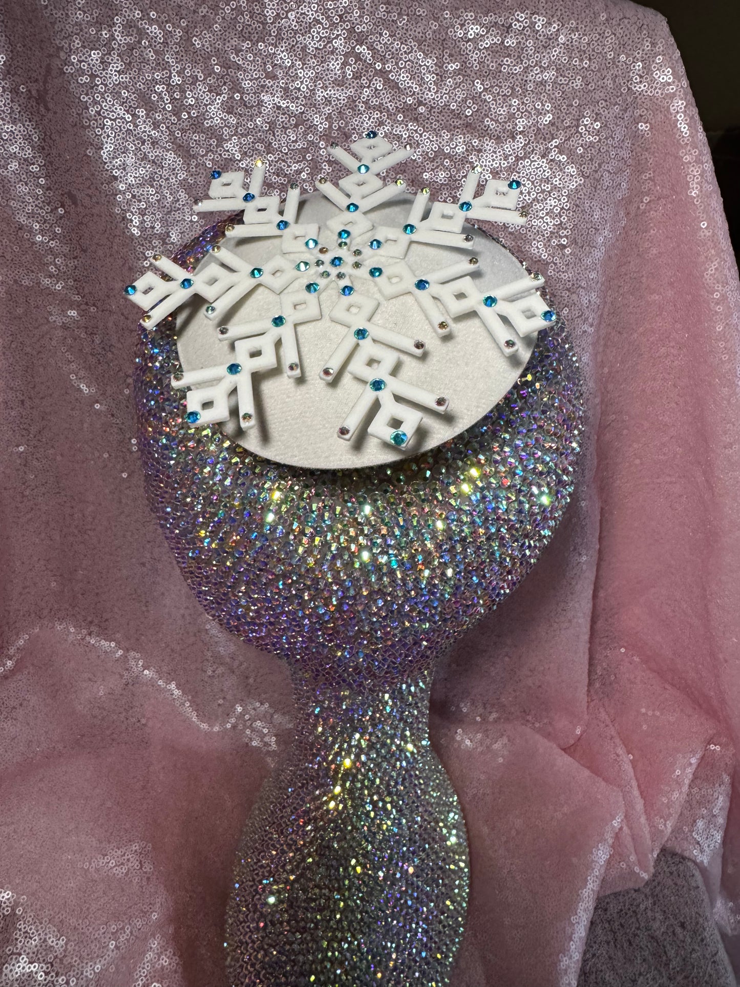 A glittering rhinestone mannequin head models the Ice Queen Fascinator by Viva La Bombshell Burlesque, featuring a silver circular base, white geometric snowflake with blue rhinestones, set against sparkly pink fabric.