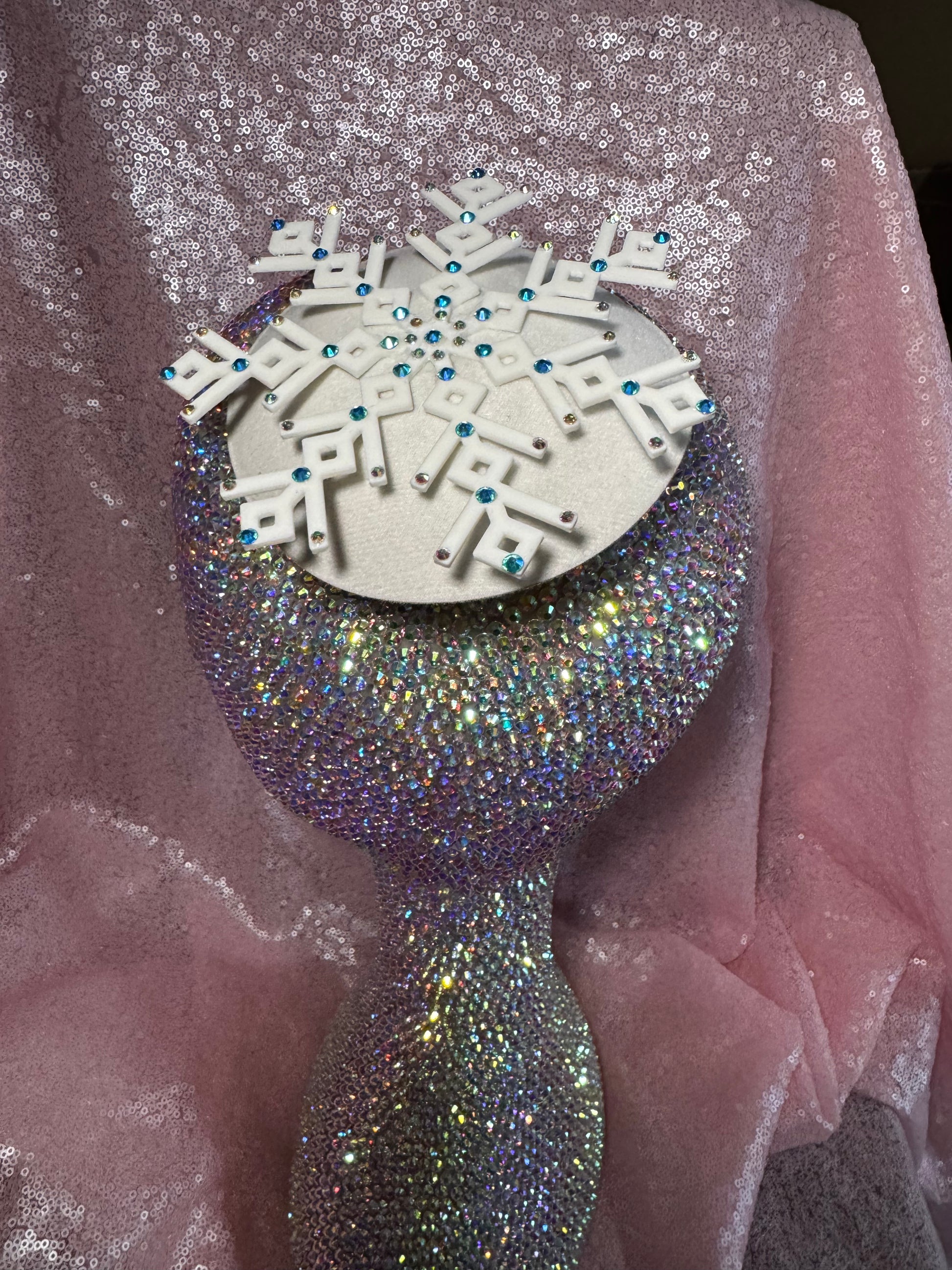 A glittering rhinestone mannequin head models the Ice Queen Fascinator by Viva La Bombshell Burlesque, featuring a silver circular base, white geometric snowflake with blue rhinestones, set against sparkly pink fabric.