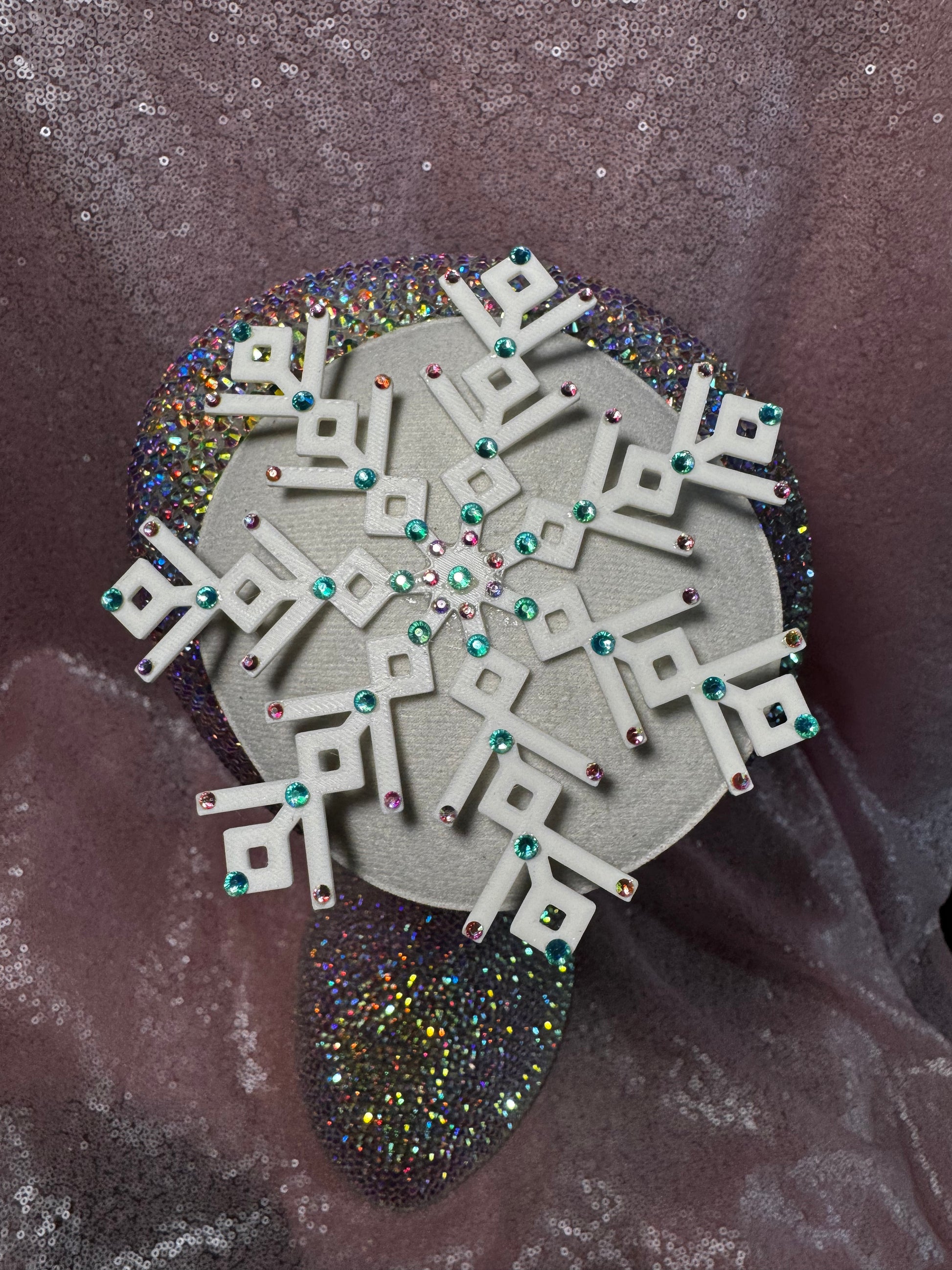 The Ice Queen Fascinator by Viva La Bombshell Burlesque, a white geometric snowflake with teal and pink gems, rests on an iridescent surface set against a shimmering pink sequin backdrop.