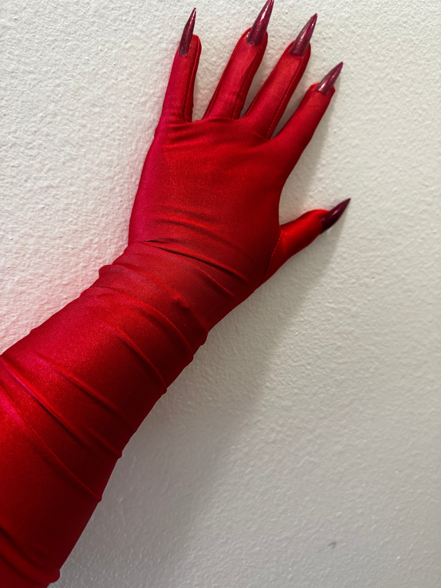 A hand wears Viva La Bombshell Burlesque's DIY Kitty Claw Gloves, featuring long, shiny, pointed dark red nails, posed against a white textured wall.