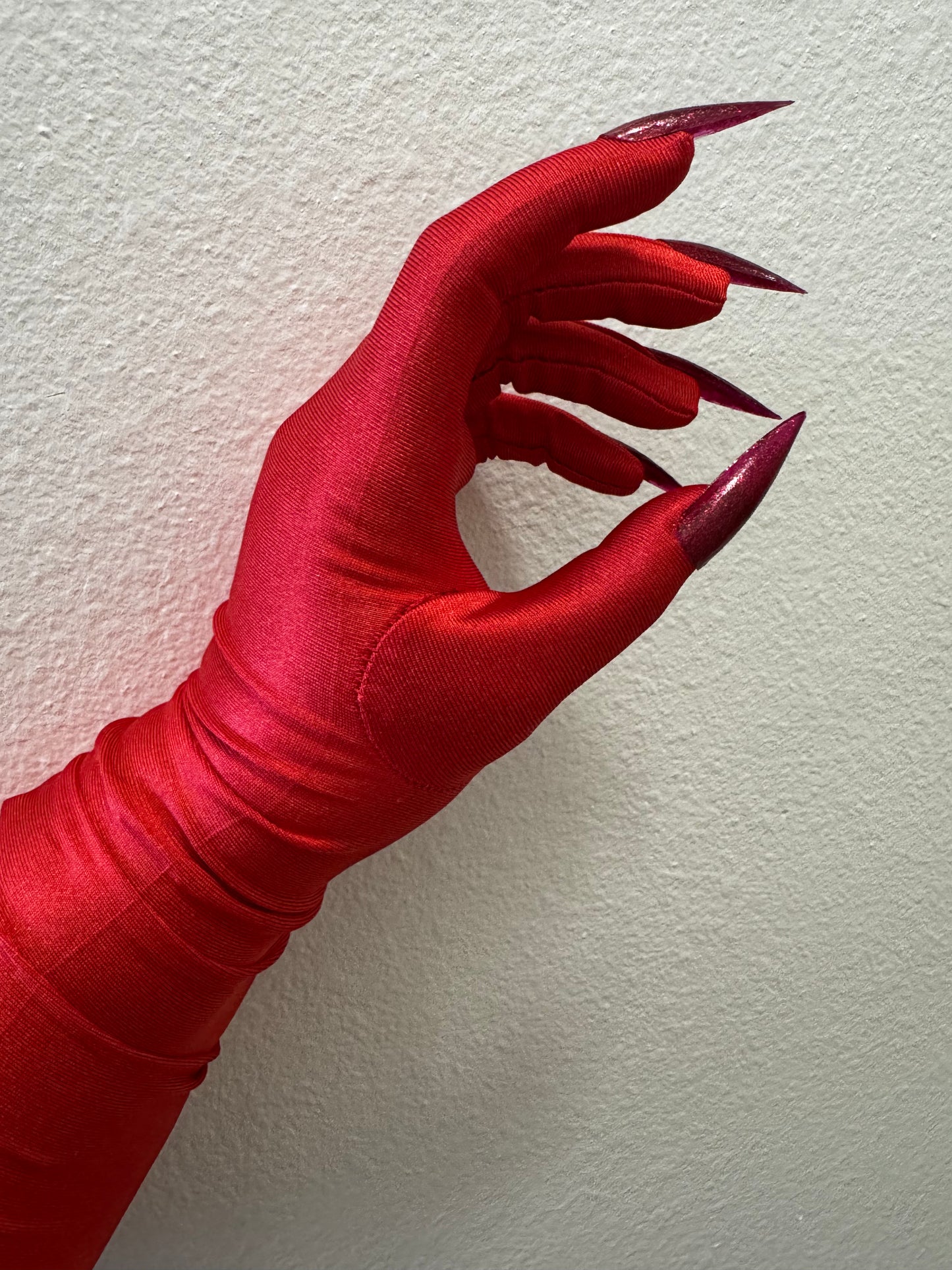 A hand wears Viva La Bombshell Burlesque’s DIY Kitty Claw Gloves—shiny red stiletto nail gloves with long, pointed nails—fingers elegantly curled against a textured beige wall.