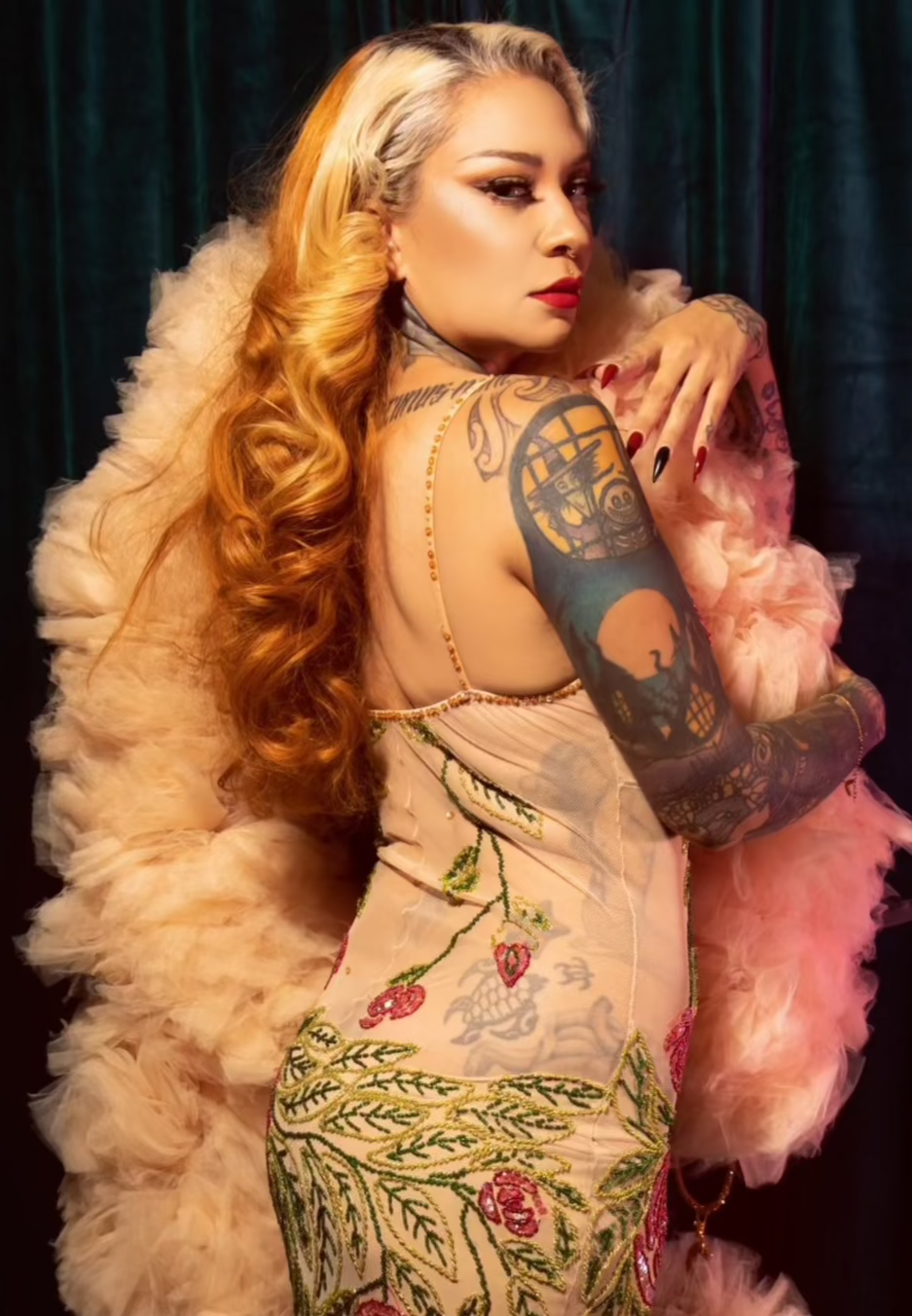 A woman with long, wavy blonde hair poses in front of a dark curtain, wearing a sheer floral dress and holding The De'Vil - Tulle Boa by Viva La Bombshell Burlesque. She sports bold makeup, colorful arm tattoos, and a delicate 3D printed charm.