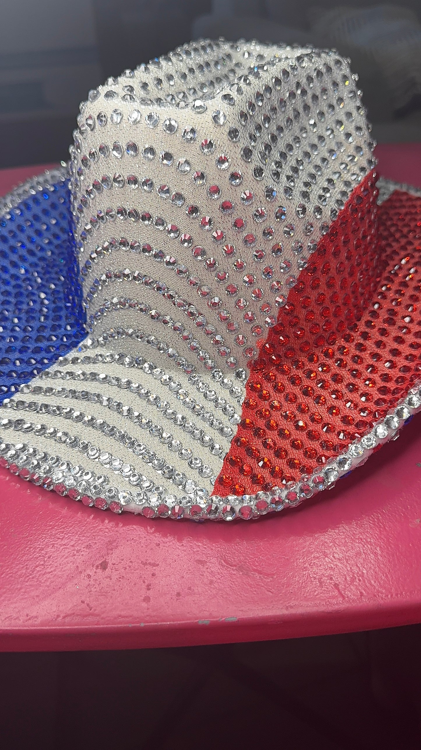 The Viva La Bombshell Burlesque Rhinestoned Cowboy Hat features silver rhinestones in red, white, and blue sections—a perfect patriotic accessory resting on a pink surface.