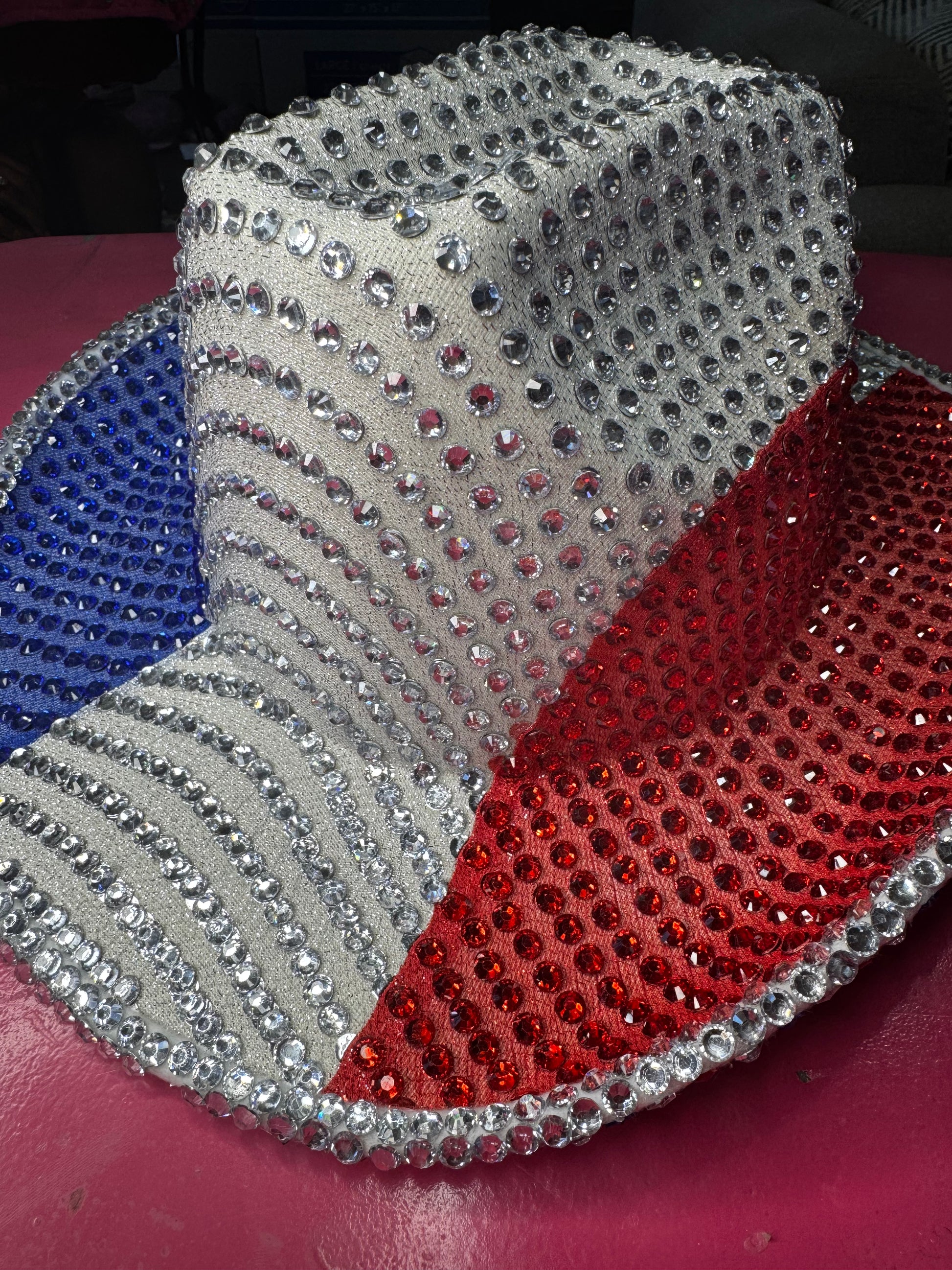 The Viva La Bombshell Burlesque Rhinestoned Cowboy Hat, adorned with silver studs and patriotic blue, white, and red sections, is displayed on a pink surface.